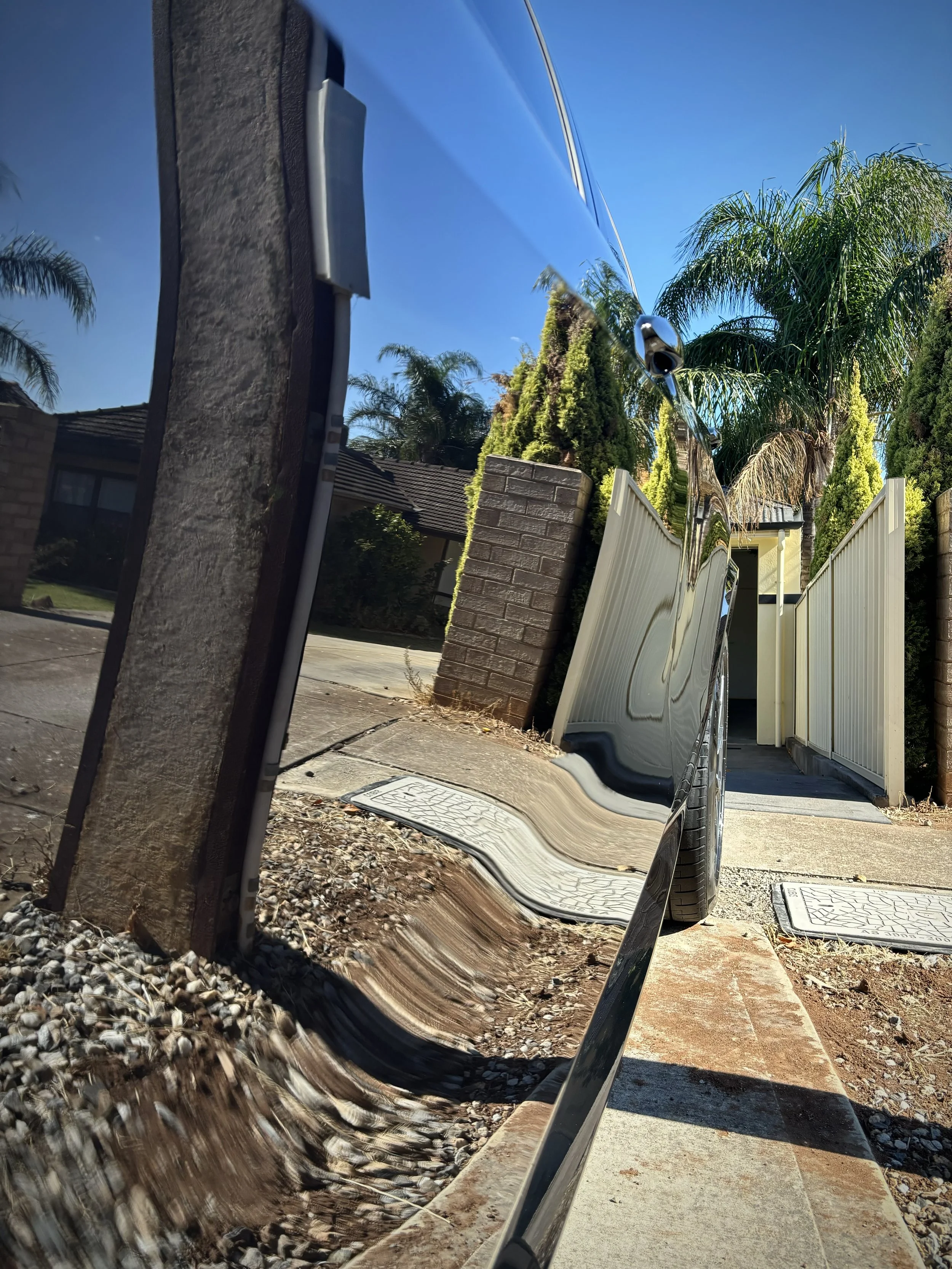 Reflected image of a house, driveway, brick wall, white fence, and palm trees in a car's shiny black side mirror under a clear blue sky.