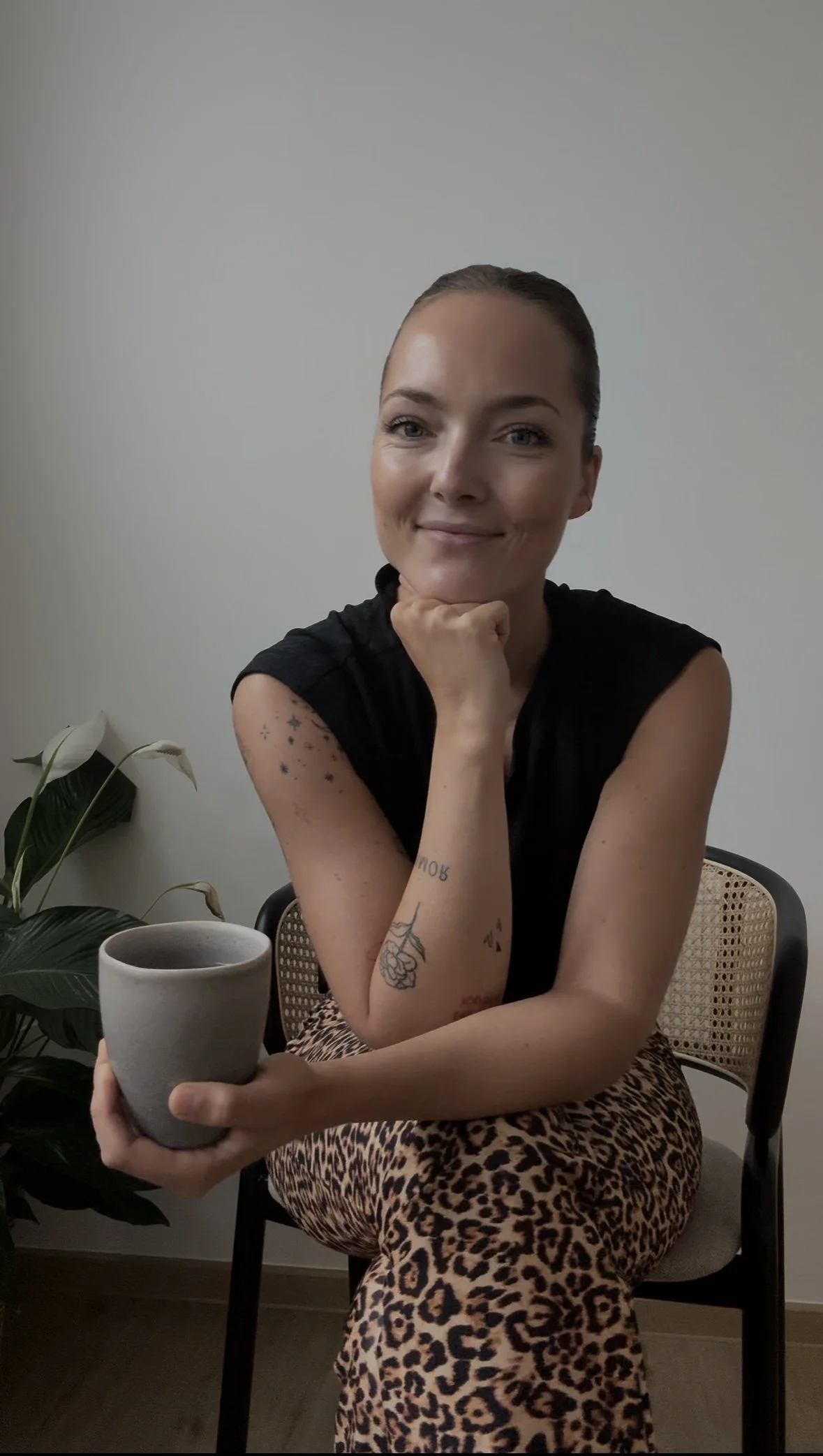 Eine lachende Frau mit Tattoos sitzt auf einem Stuhl, hält eine Tasse und trägt ein schwarzes Top und Leopard-Print-Hose vor einem weißen Hintergrund.