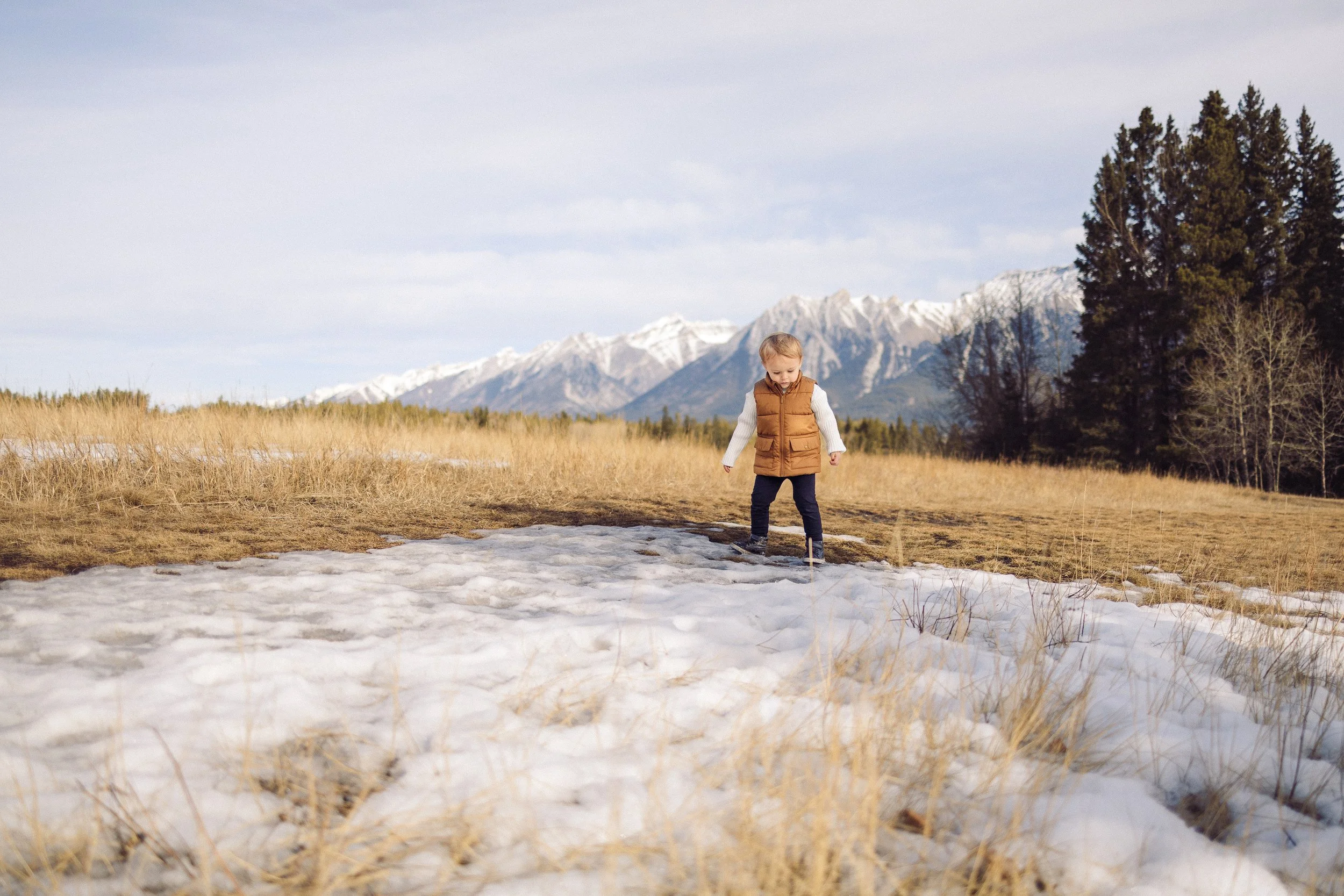 A young child walks on snow and dried grass with snow-capped mountains, trees, and a cloudy sky in the background in Canmore.