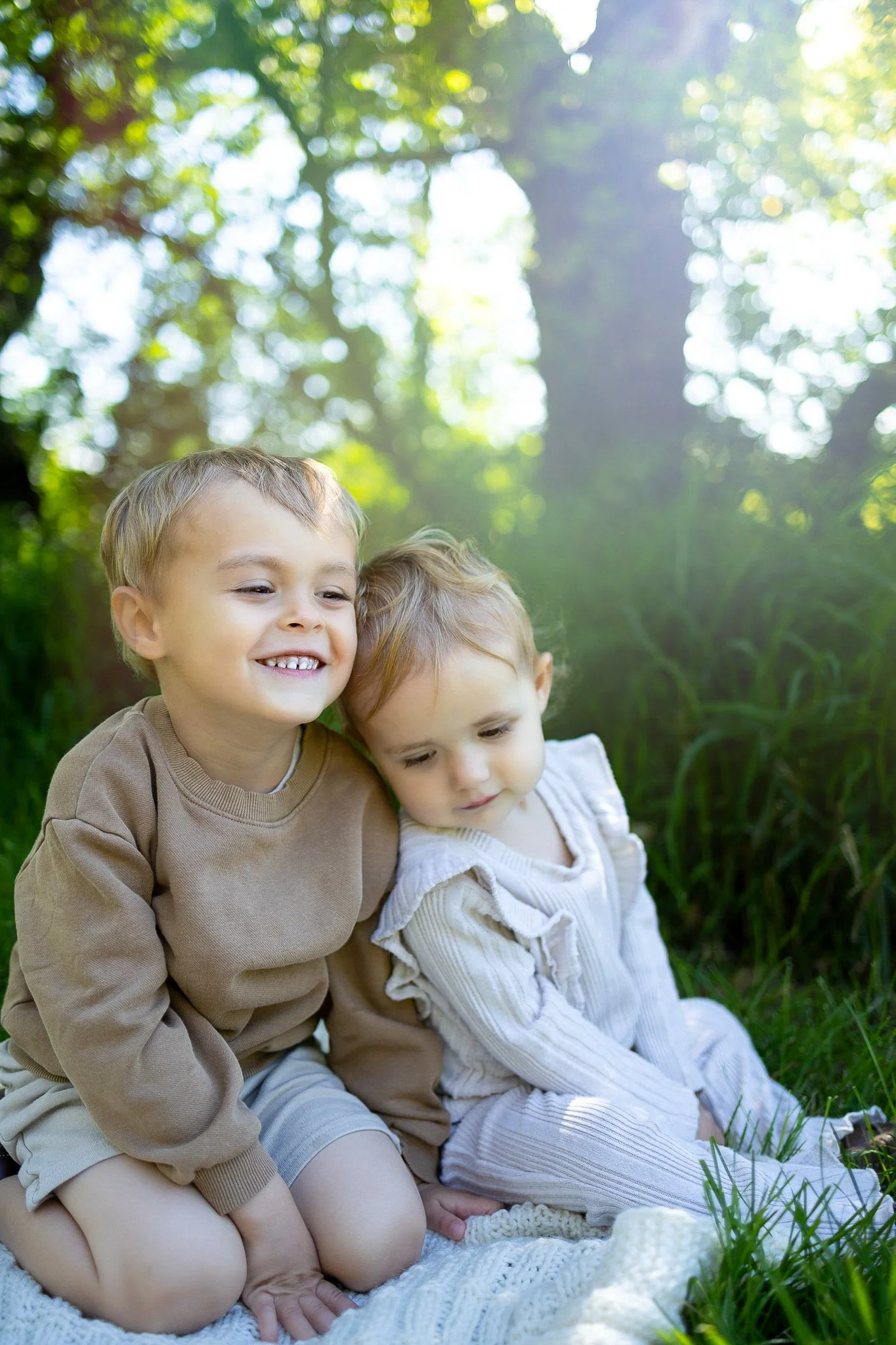 Two young children siblings sitting outdoors on a blanket in a grassy area with trees in Fish Creek Provincial Park.