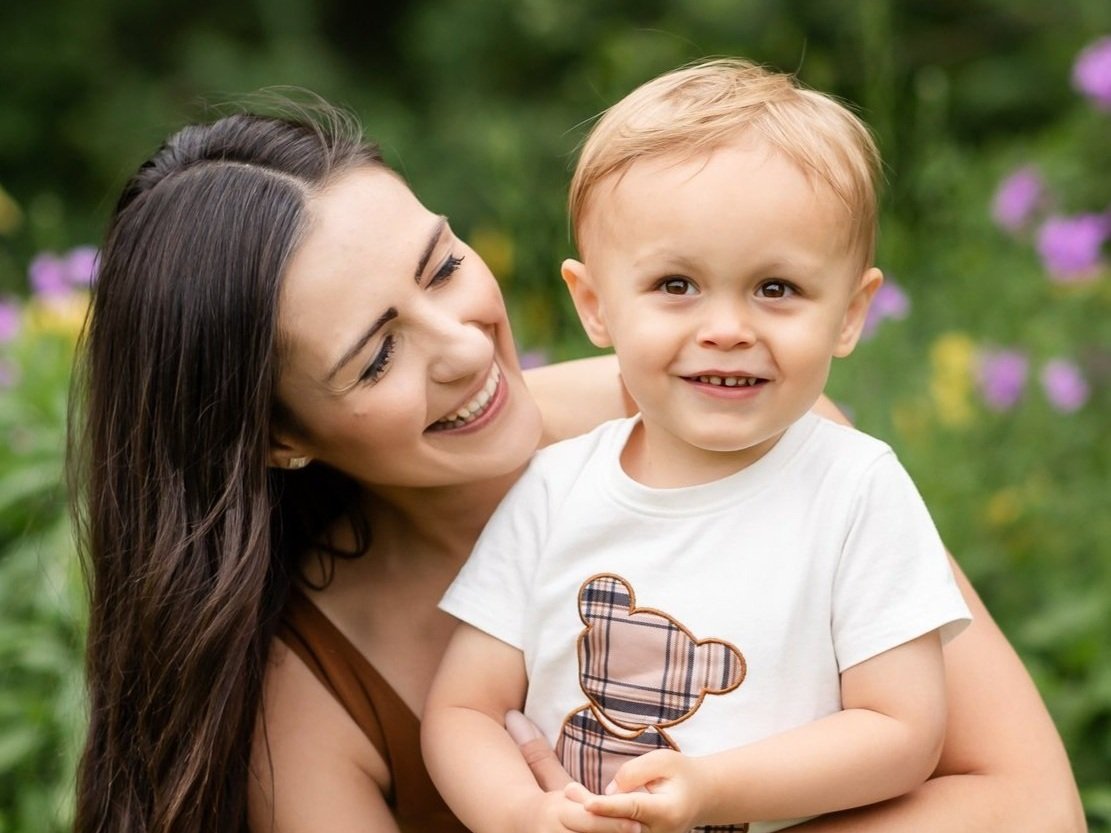 A woman smiling and holding a young boy outdoors with greenery and purple flowers in the background.