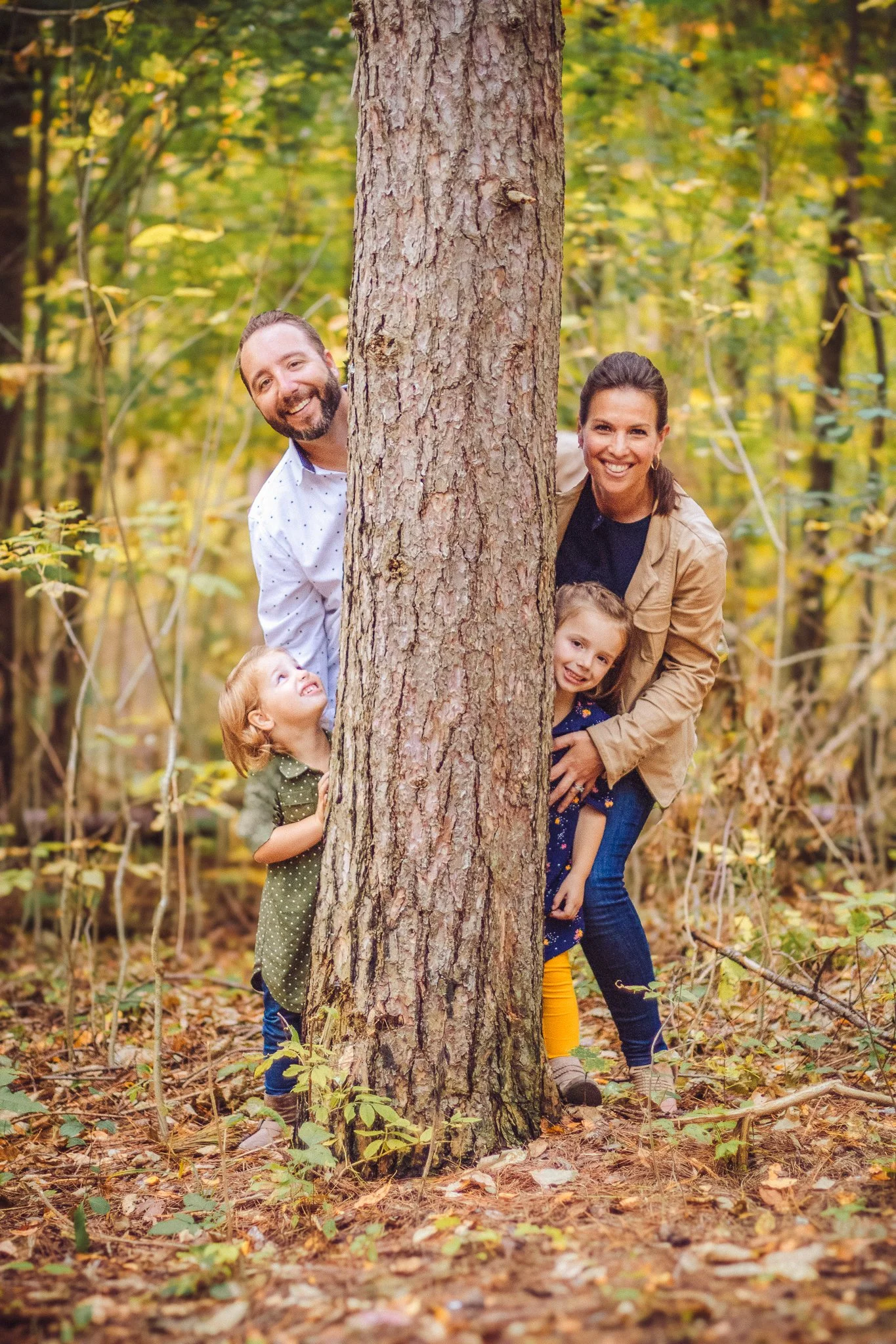 A family of four peeking out from behind a large tree trunk in a forest during fall in Calgary for a photography session.