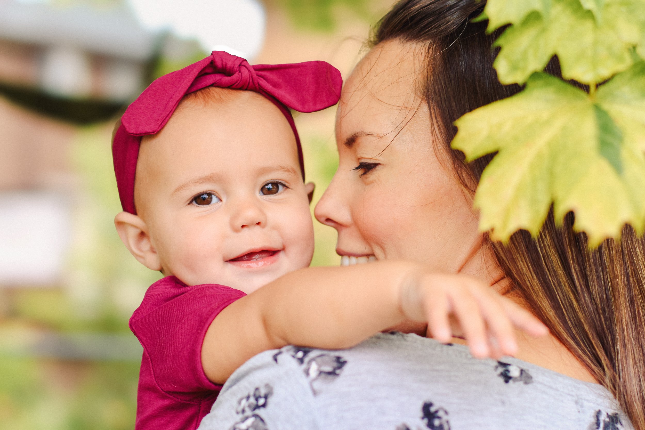 A woman holding a smiling baby girl outdoors, with greenery and trees partially visible in the background in Calgary.