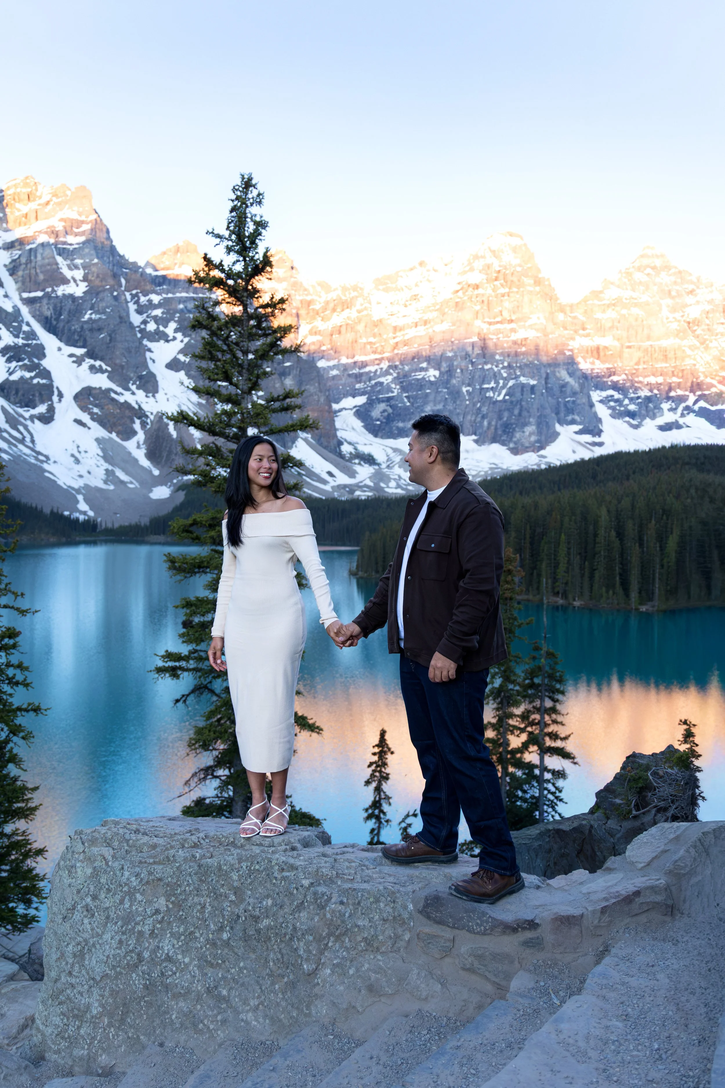 A couple holding hands and facing each other on a large rock in a mountainous landscape with snow-capped peaks, a lake, and a few trees, during sunrise at Moraine Lake in Banff for a couples photo session.