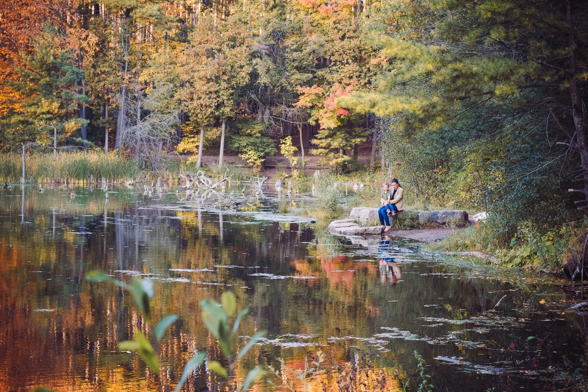 A woman and a child sit on a large rock by the edge of a river in a forested area during autumn, with trees displaying colorful fall foliage.