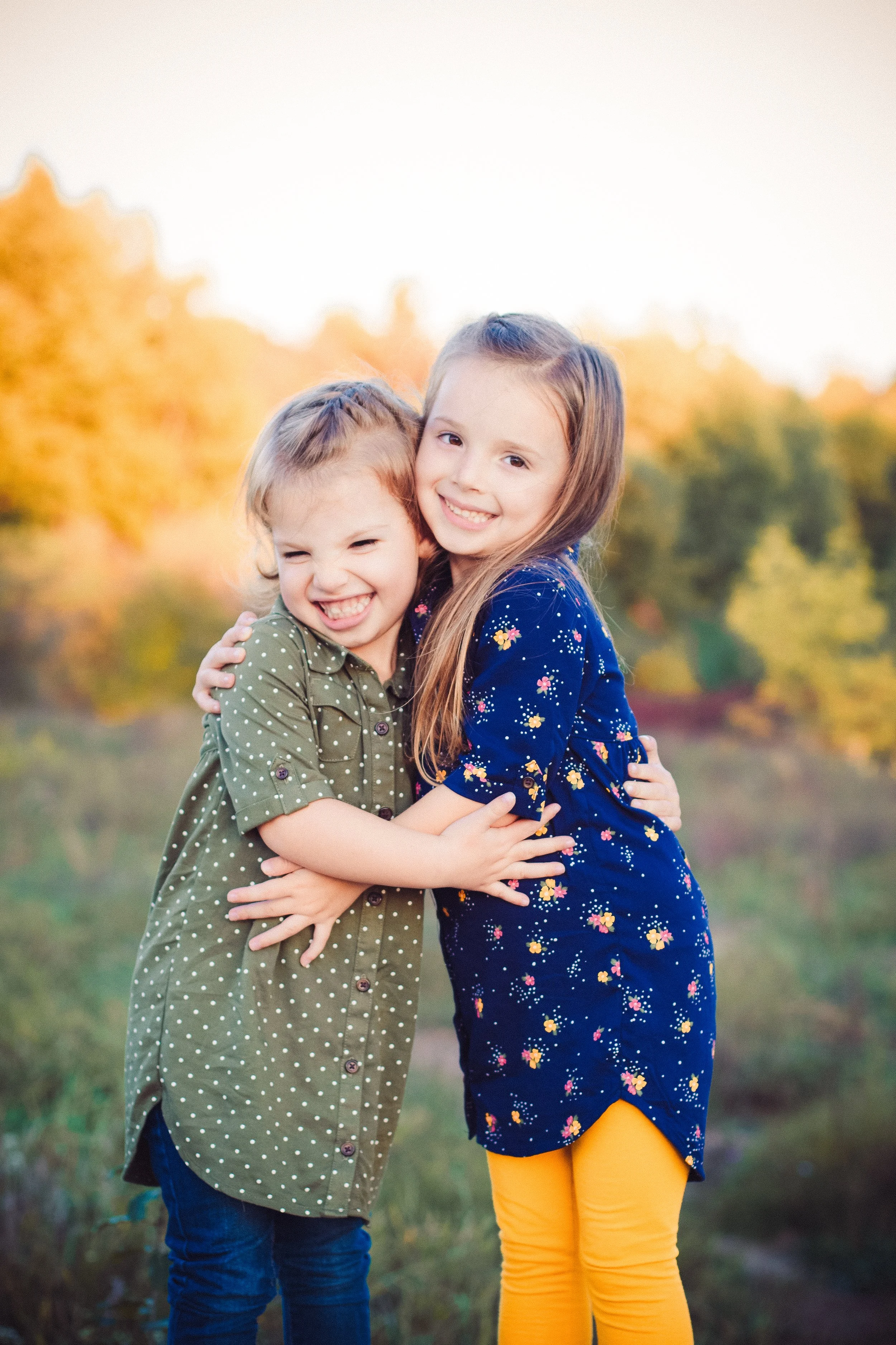 Two young girls hugging outdoors during autumn with colorful trees in the background.