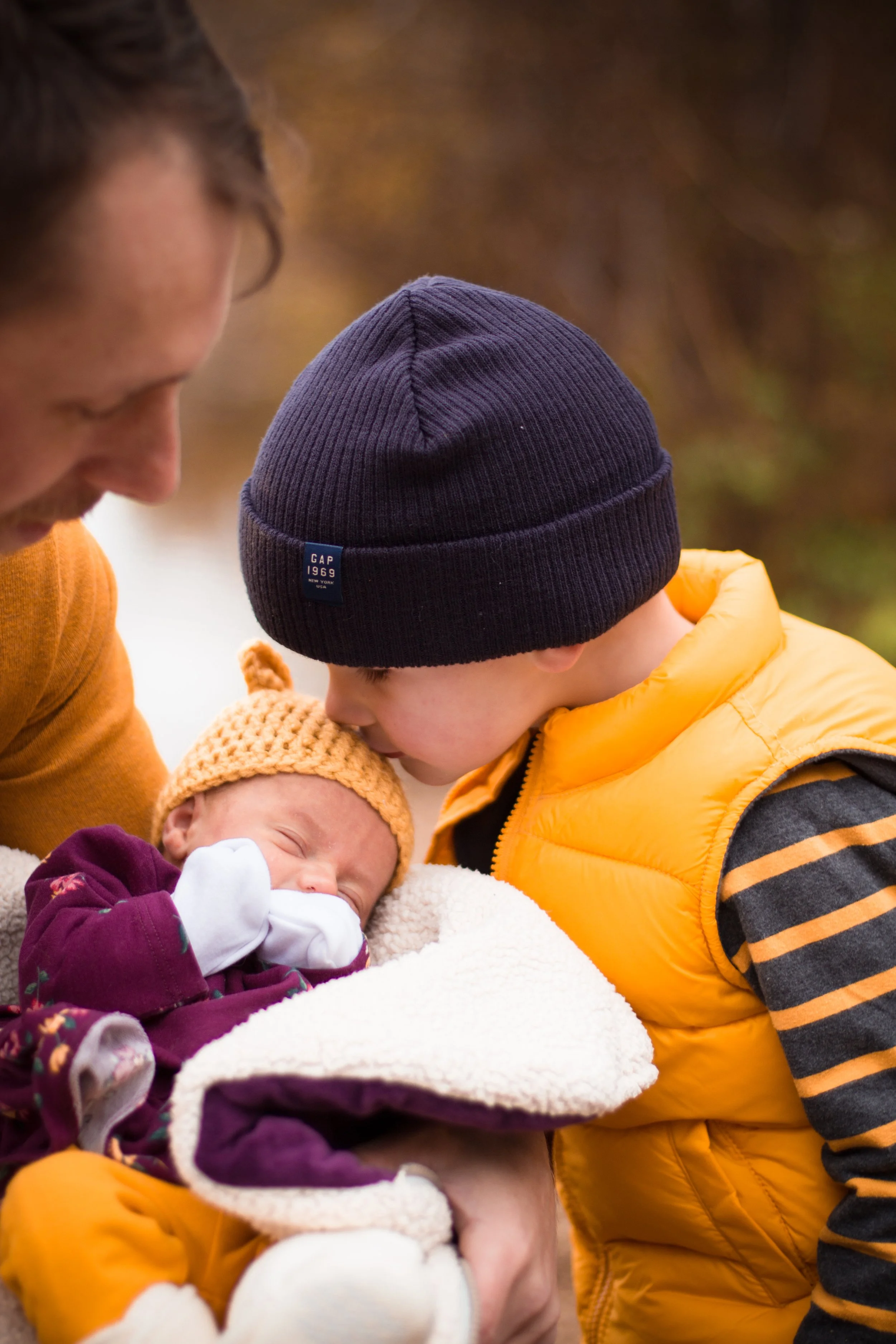 A young boy in a navy beanie and yellow puffer vest kisses a newborn baby wrapped in a fleece blanket, with a man watching them outdoors in Calgary.