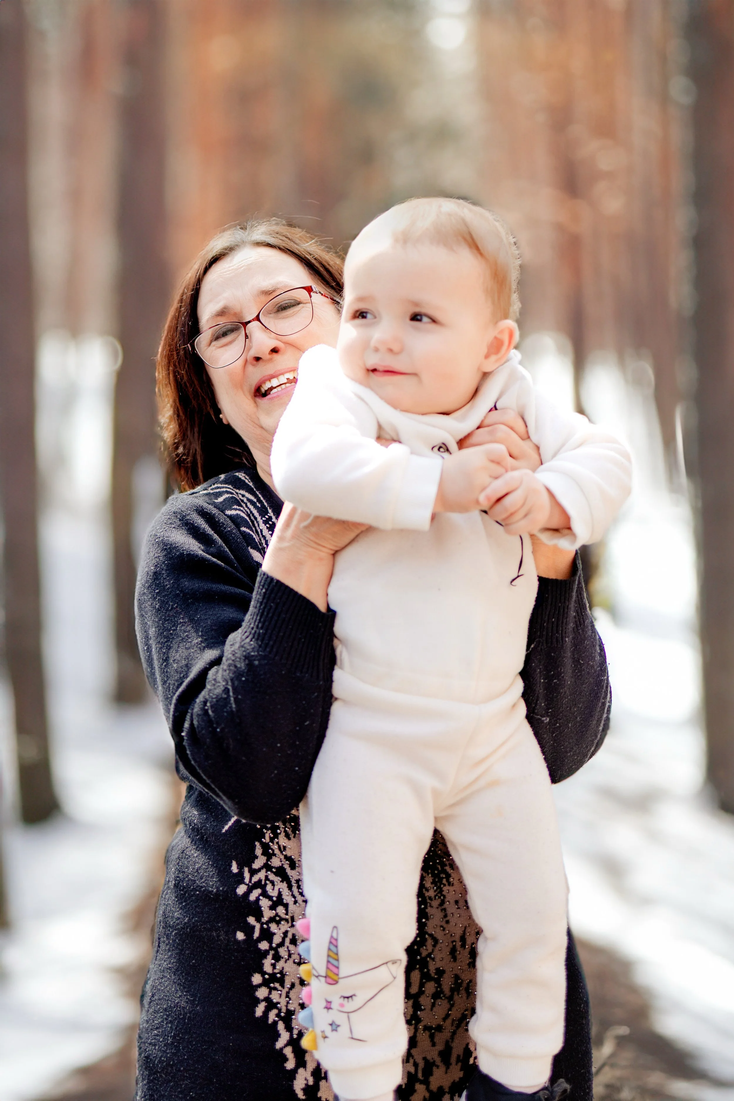 A grandmother with glasses holding a young child in a forest with tall trees, some snow on the ground, in Calgary.