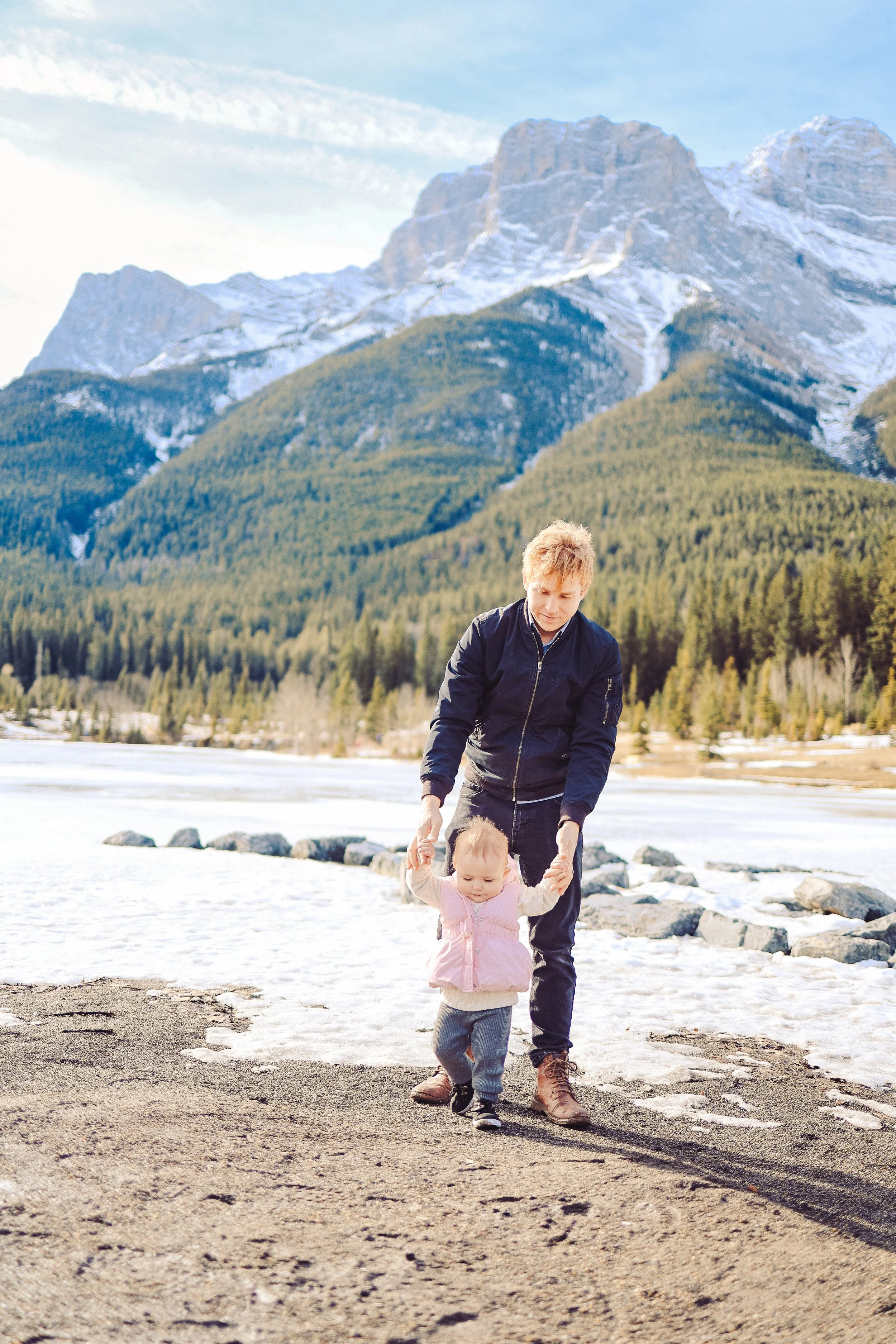 A father helping their baby walk on a dirt path near a partially frozen lake with mountains and a forest in the background in Canmore.