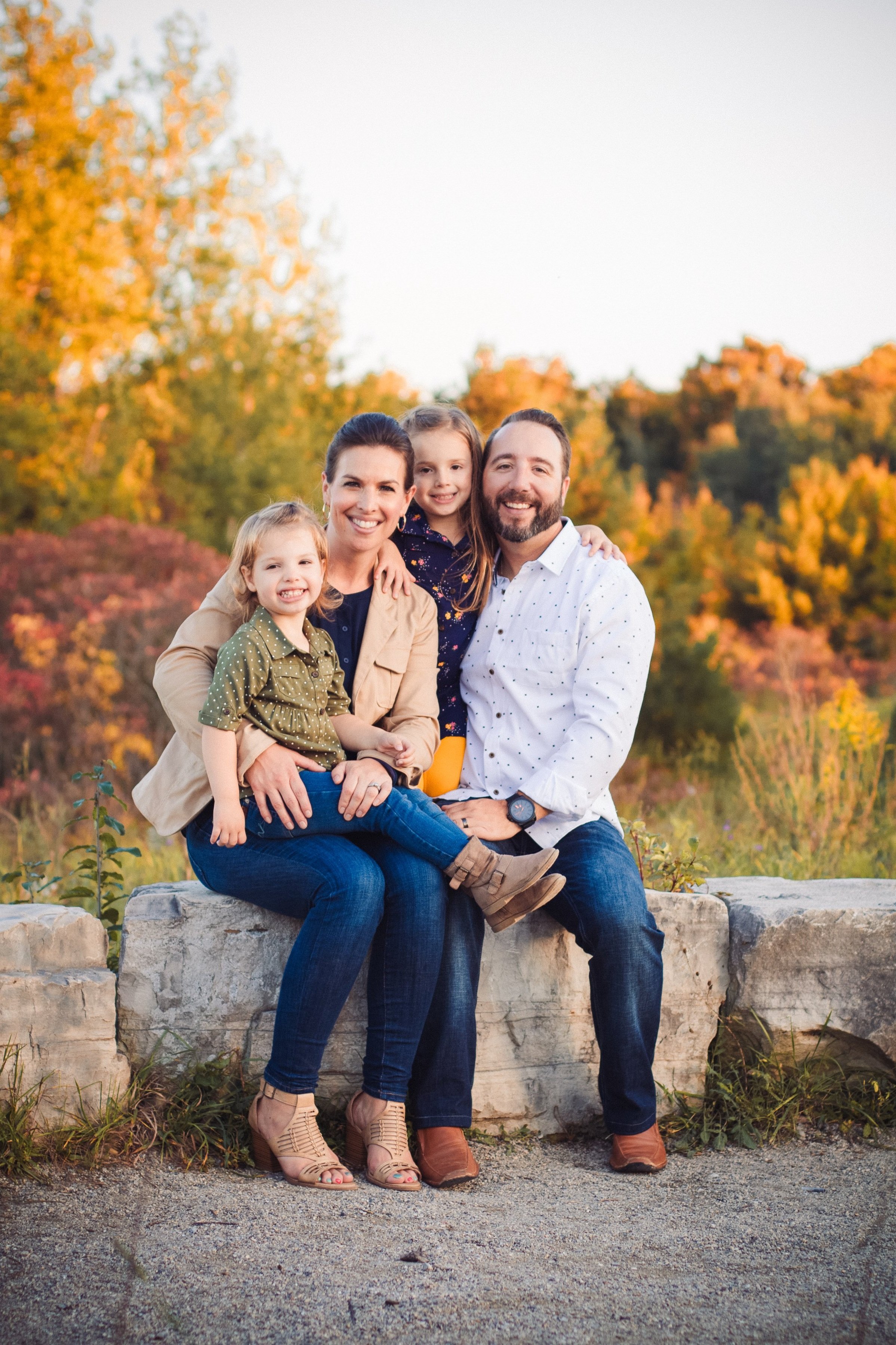 A family of four, including a mother, father, and two young daughters, sitting on a stone border in Calgary outdoors during autumn with vibrant fall foliage in the background, smiling at the camera.