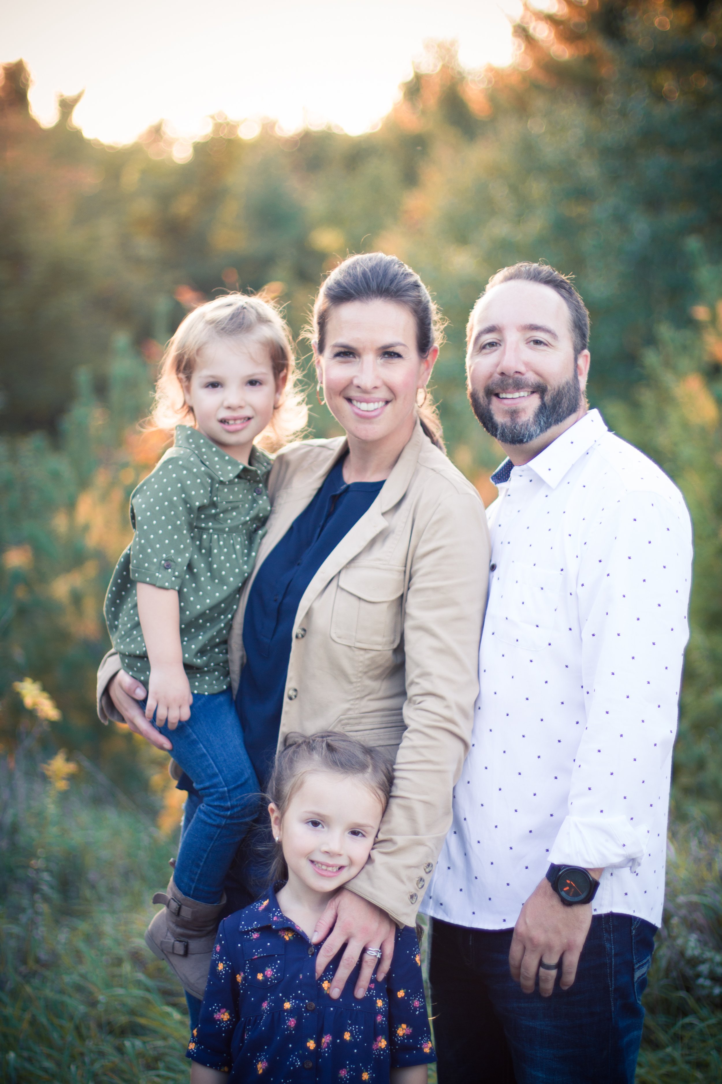 A family of four outdoors in a park during sunset, including a mother, father, and two young daughters, smiling at the camera.