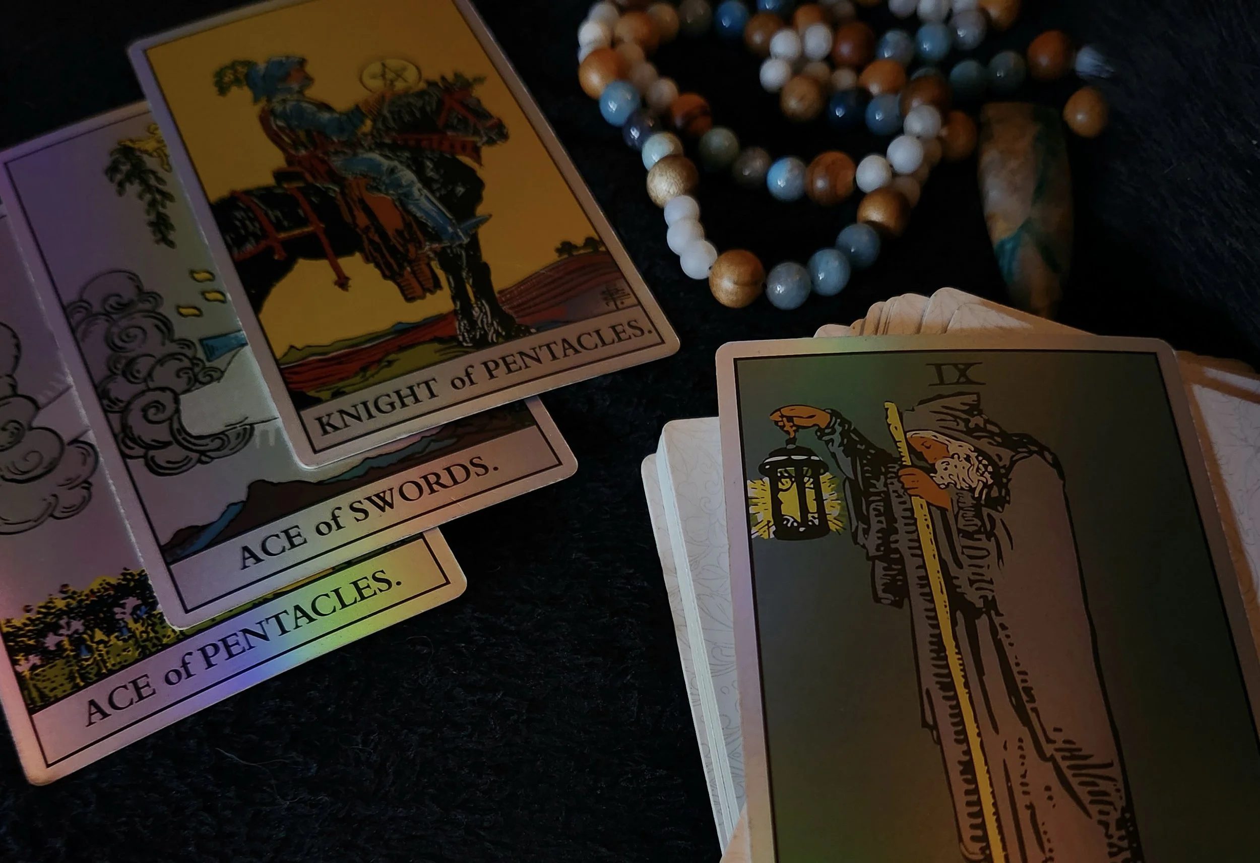3 tarot cards sit on top of each other in the leftmost corner, a necklace of blue & white crystal beads with wooden beads interchangeable lay in the rightmost corner, The Hermit tarot card covers the rest of the image