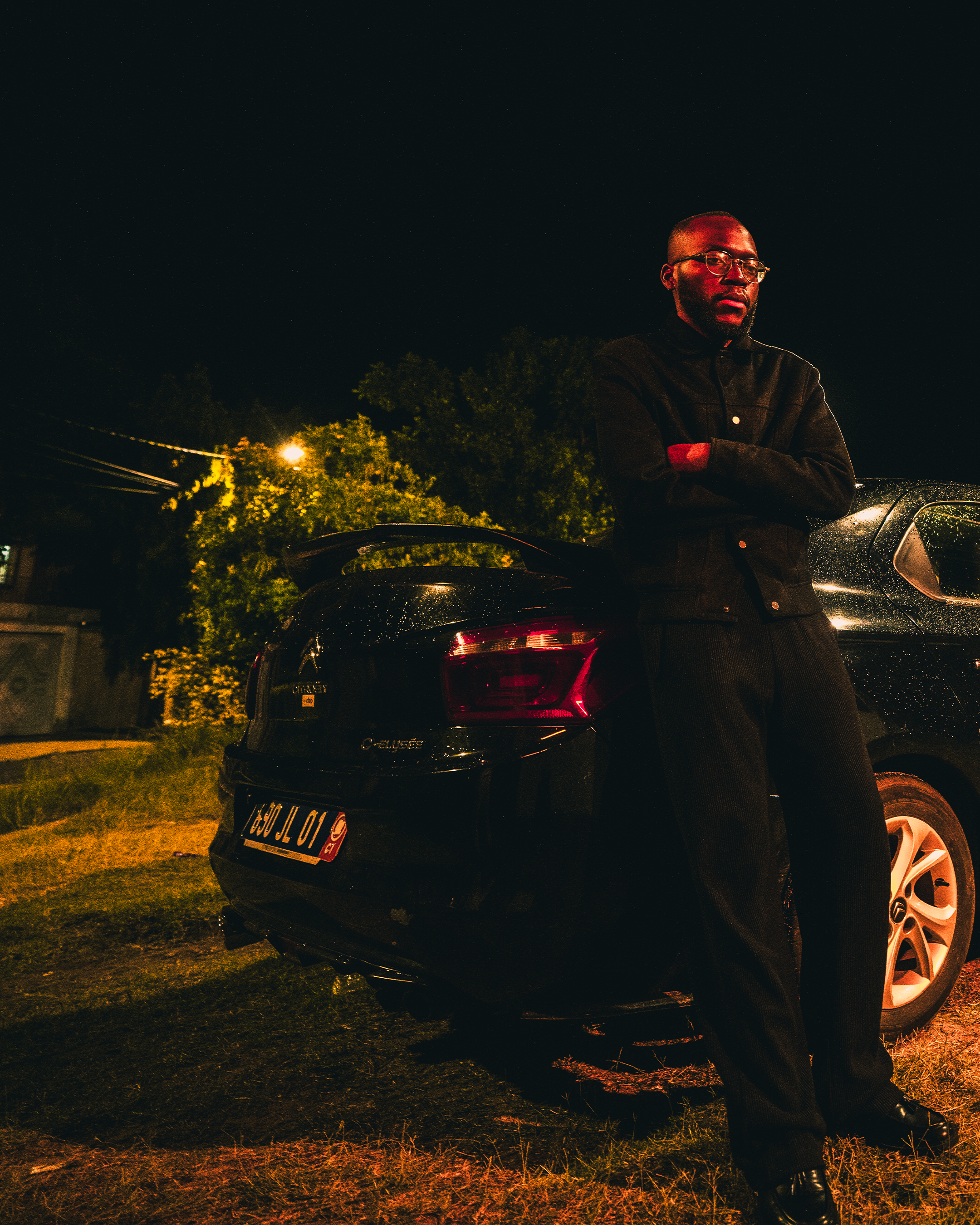 Un homme debout à côté d'une voiture noire, la nuit, avec un ciel étoilé. Il porte des lunettes et une veste noire, ses bras croisés.