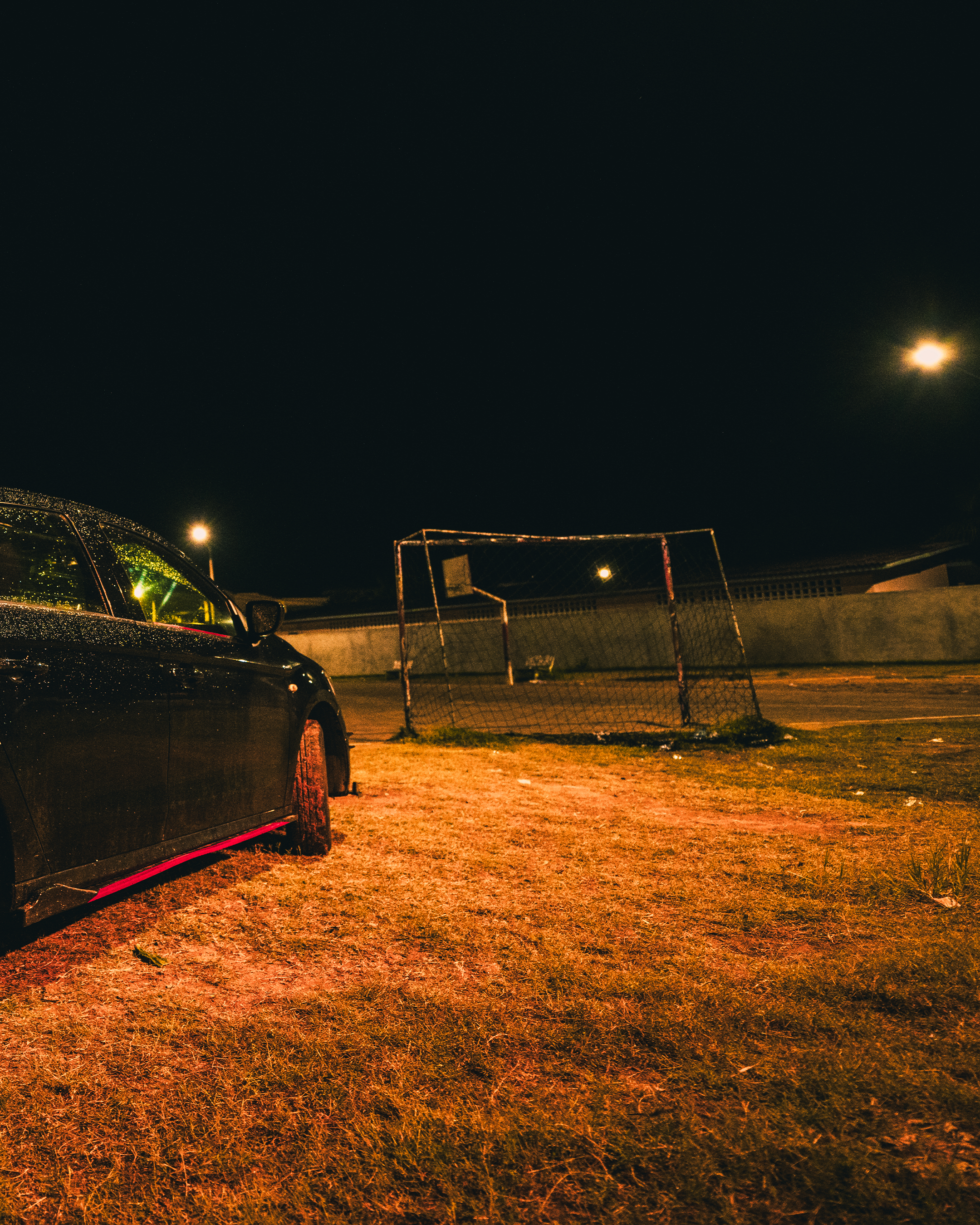 Un terrain de football vide la nuit avec un but rouillé, une voiture noire garée à gauche, et des lampadaires illuminant le sol et le ciel noir.