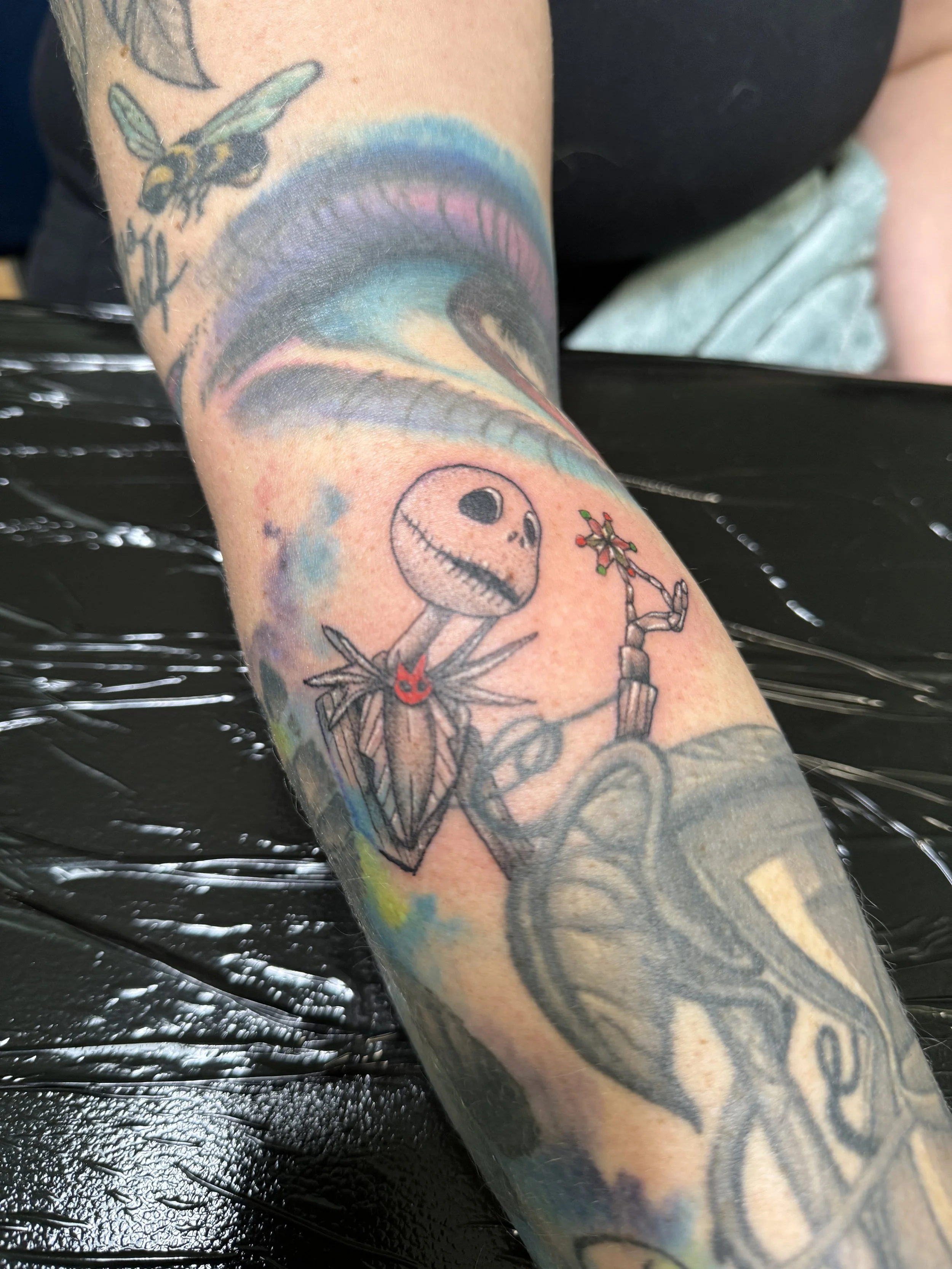 Tattoo of Jack Skellington holding a rose in his hand, with colorful rainbow background and other tattoo designs visible.