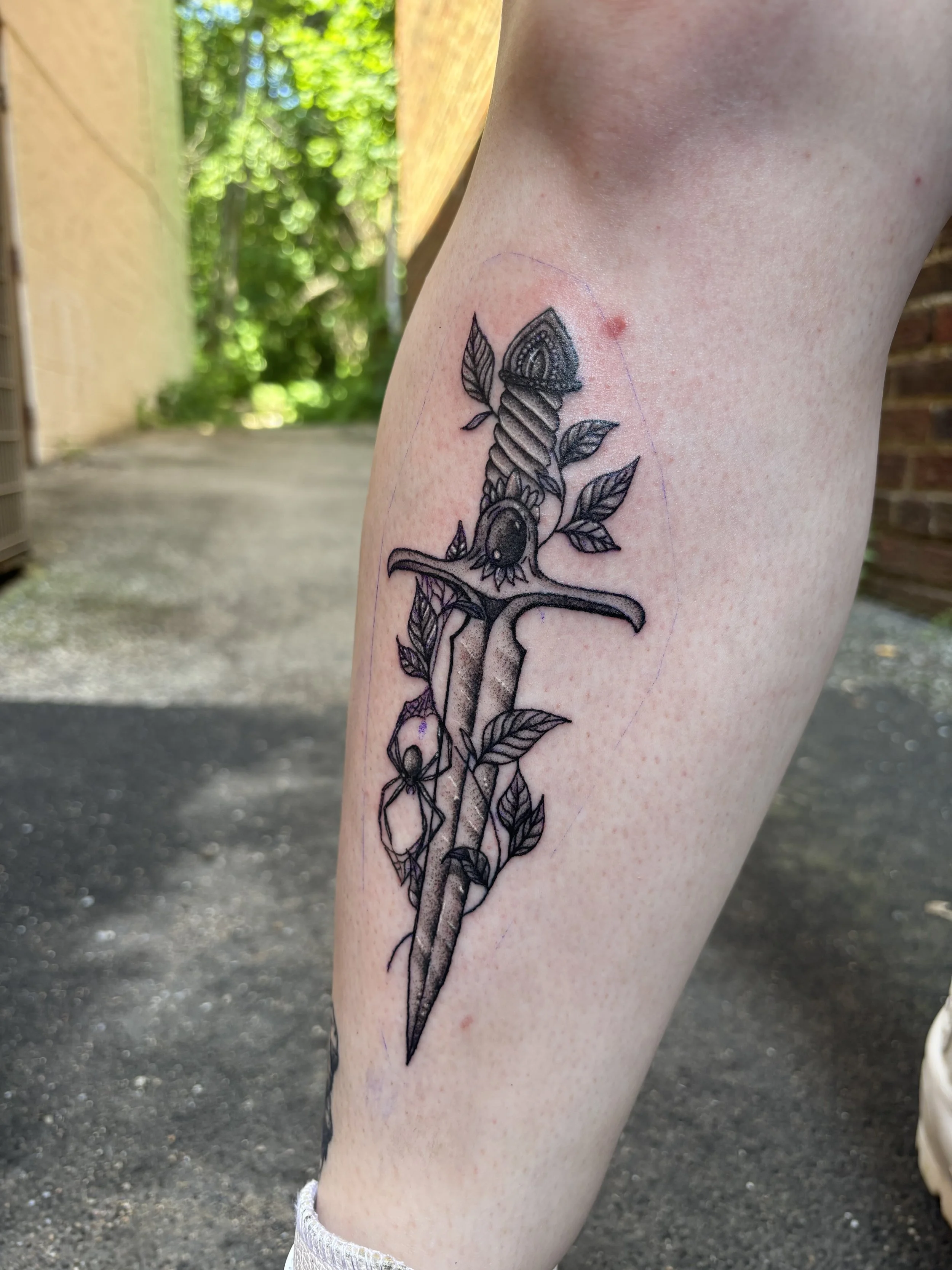 A black ink tattoo of a sword entwined with leaves and a butterfly on a person's calf.