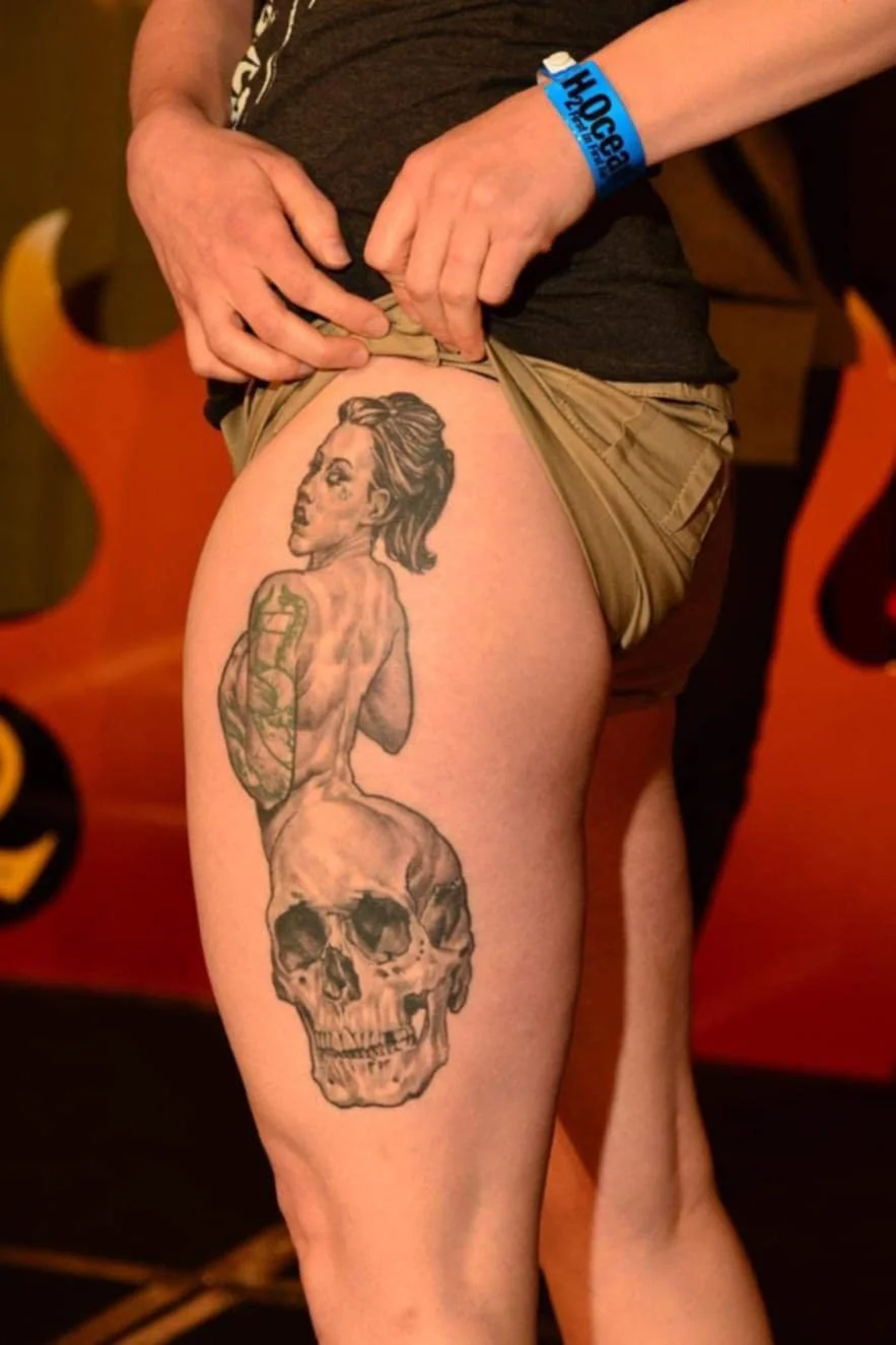 A person's thigh features a large tattoo of a woman with short hair and a skull, with the woman partially covering the skull's top.