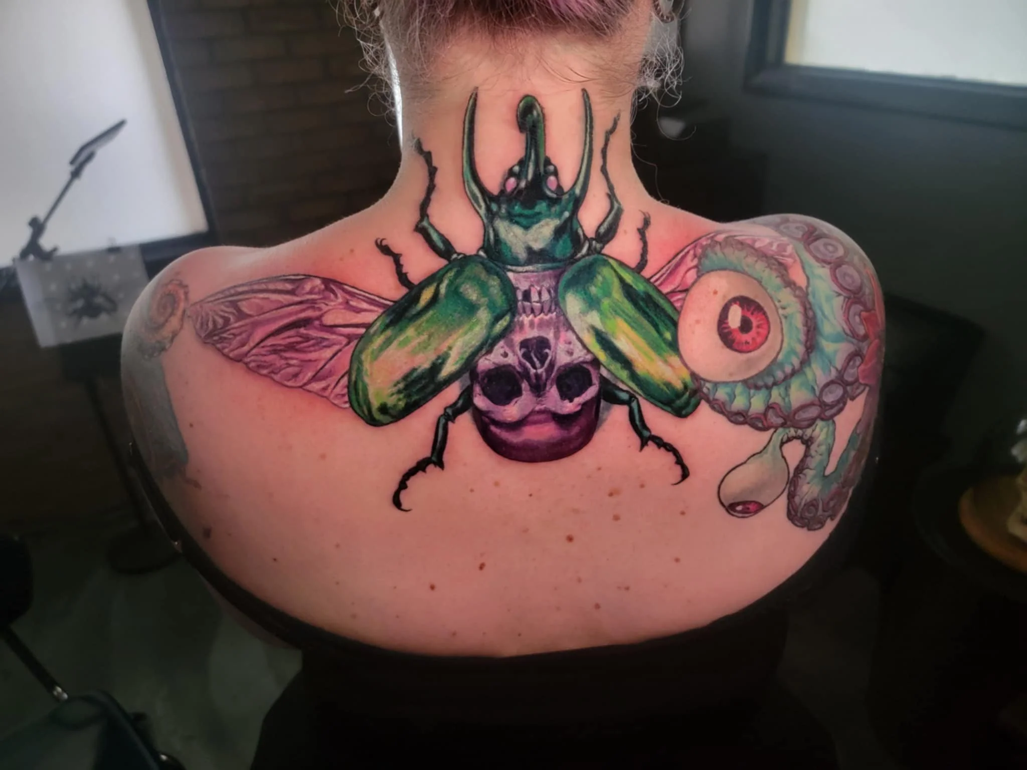 Colorful tattoo of a large insect with a beetle body, green wings, and a head resembling a stag beetle, on a woman's upper back and shoulders, featuring an eye and an octopus tentacle.