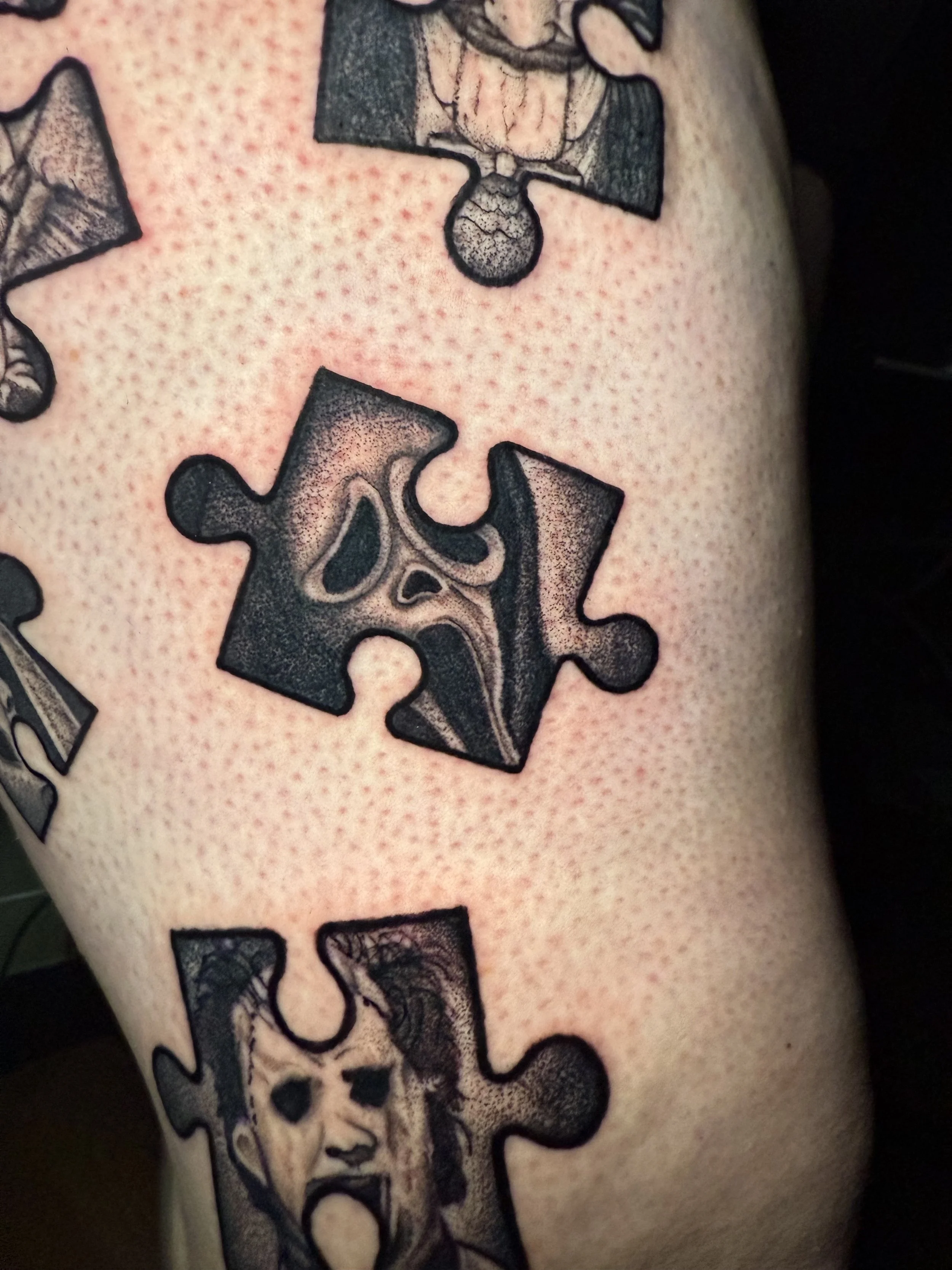 Close-up of a tattoo on skin featuring puzzle pieces with artistic faces, one of which appears to be a skull, and intricate shading, with a dotted background.