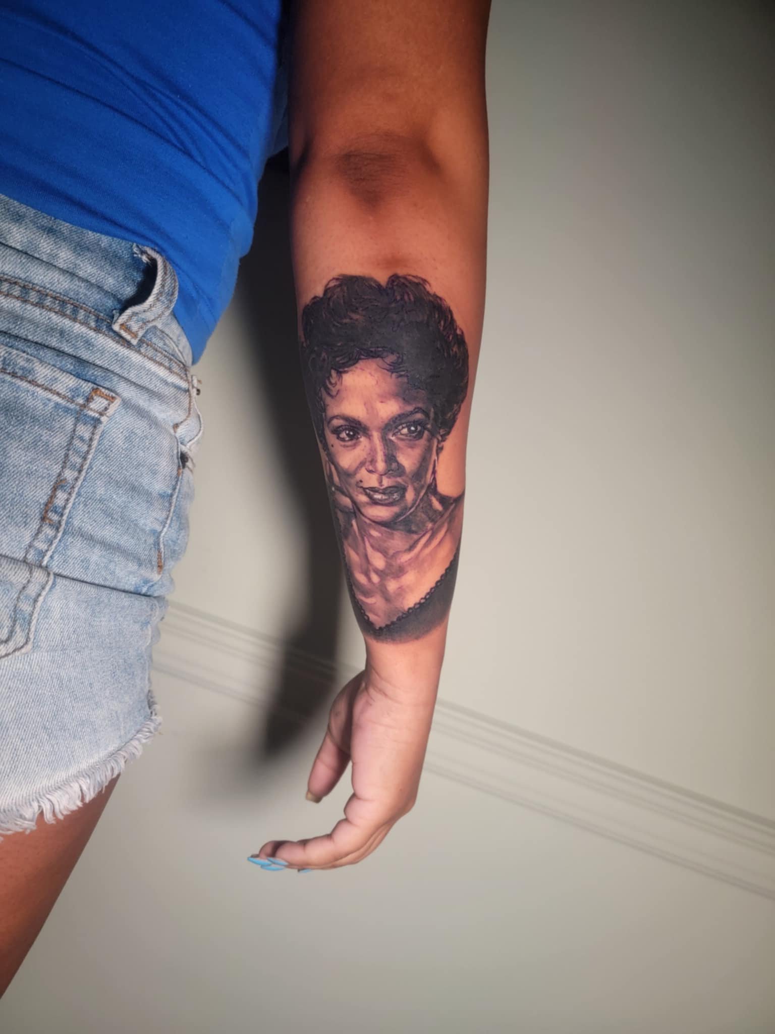A person's arm with a realistic black and gray tattoo of a woman's face, wearing a blue shirt and light blue denim shorts.