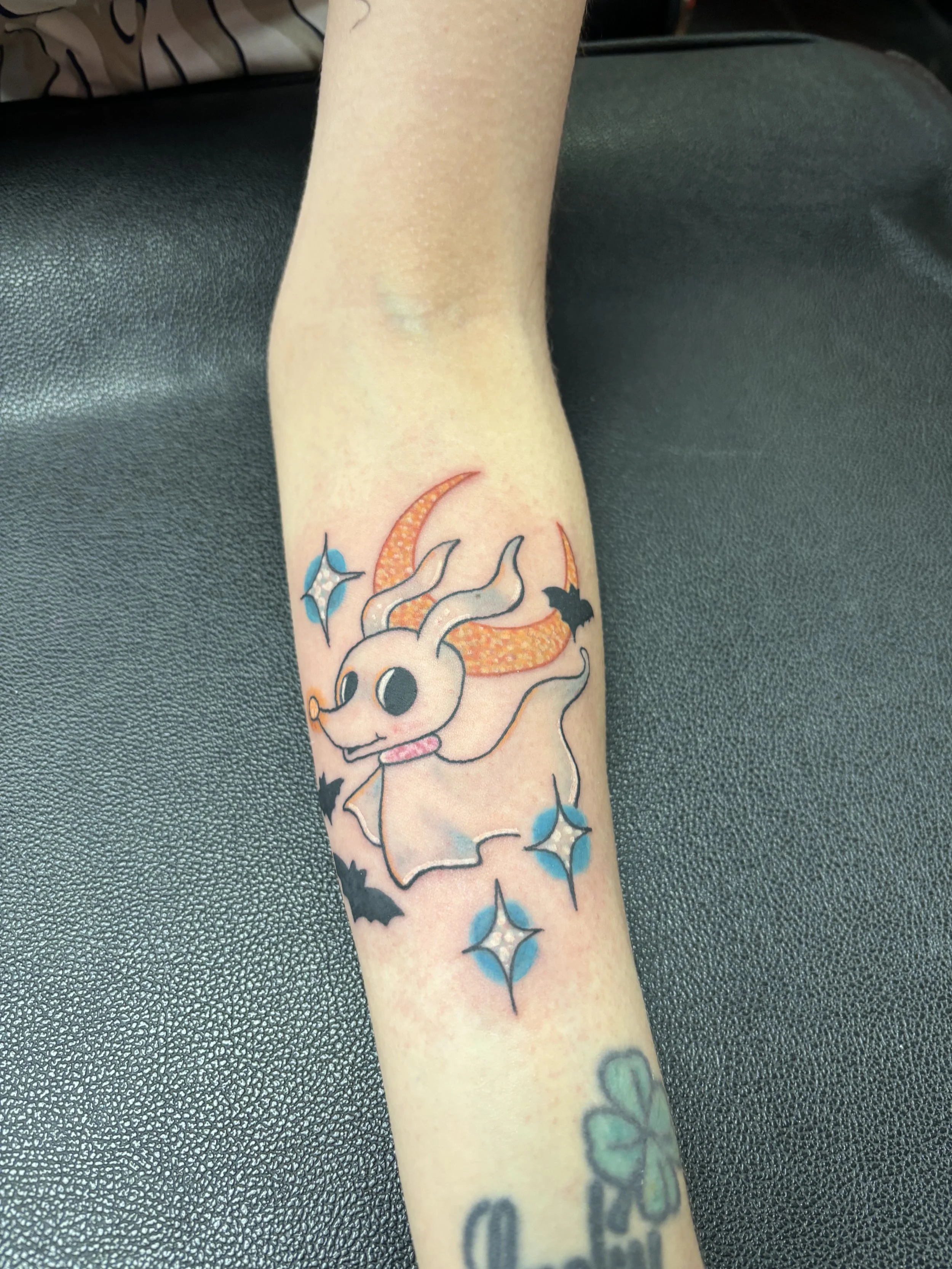 Tattoo of a cartoon-style goat with orange horns, surrounded by blue and black star shapes and small bats, on a person's forearm.