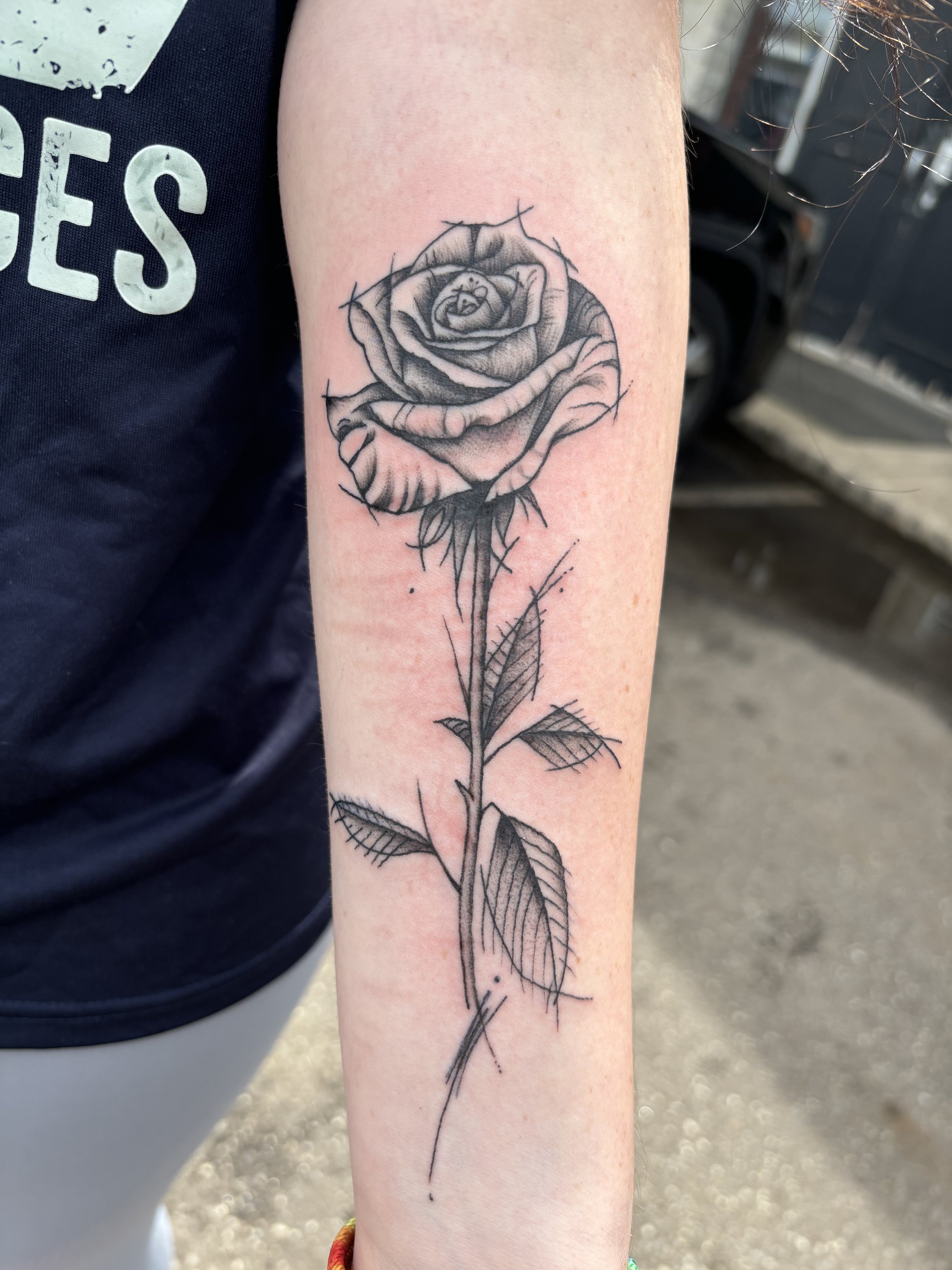 Black and gray tattoo of a rose with thorns and leaves on an inner forearm.