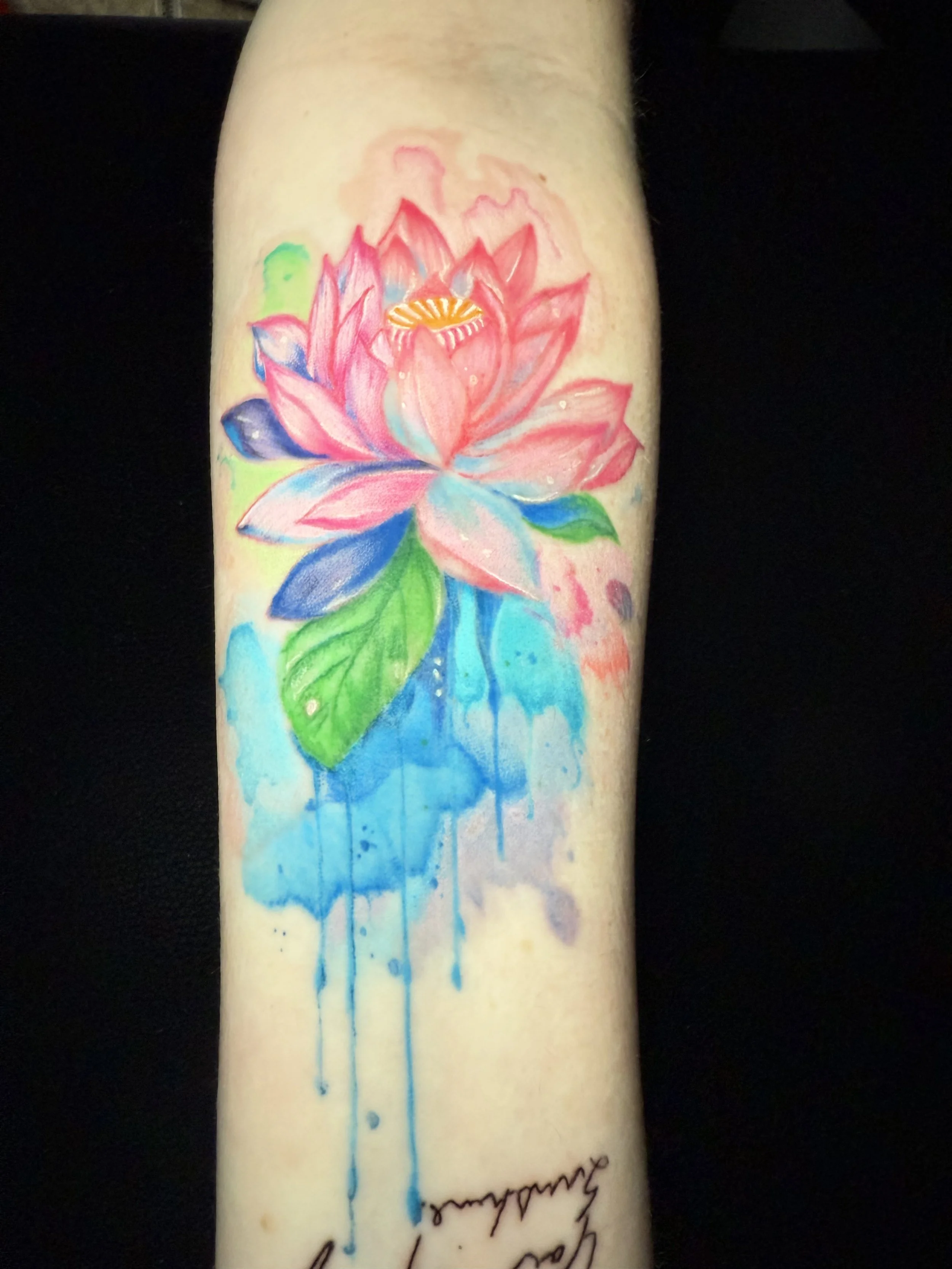 Colorful watercolor tattoo of a pink and blue lotus flower with green leaves and blue paint drips, with a signature at the bottom.