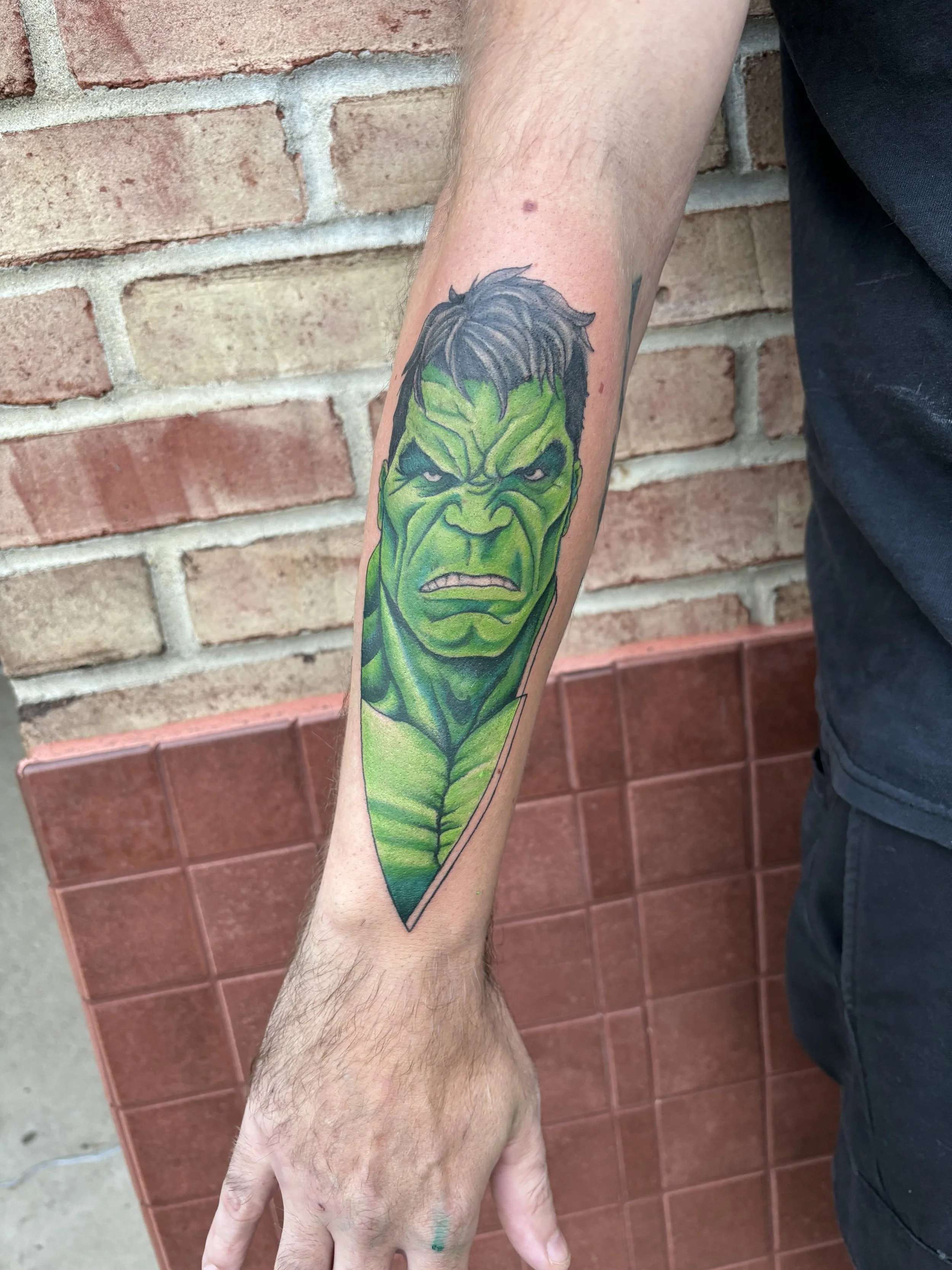 Tattoo of the Hulk's face on a person's forearm, with green skin and angry expression, set against a brick wall background.