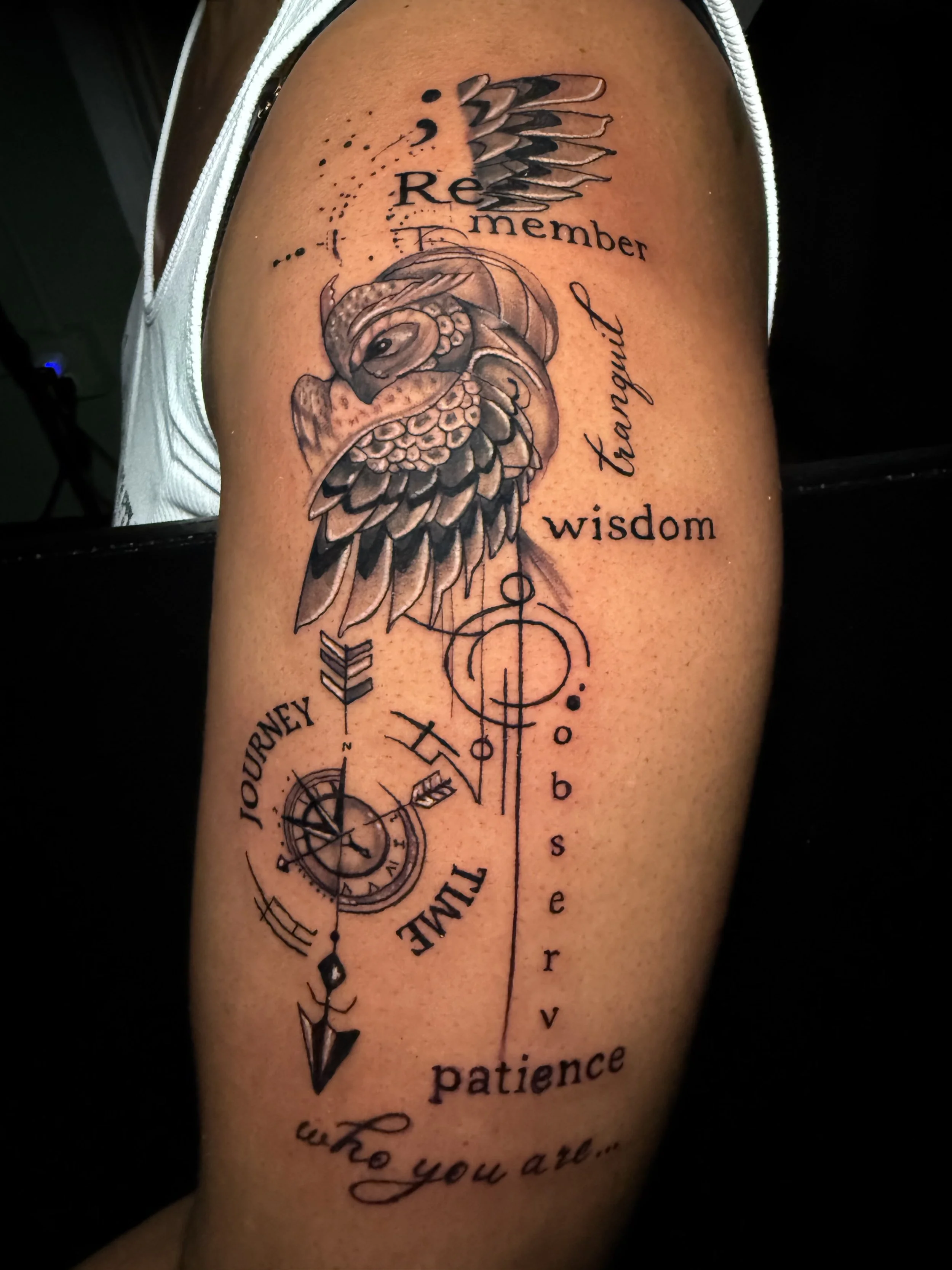 A tattoo on a person's upper arm featuring an owl, feathers, a compass, and scriptures about journey, wisdom, patience, and remembrance.