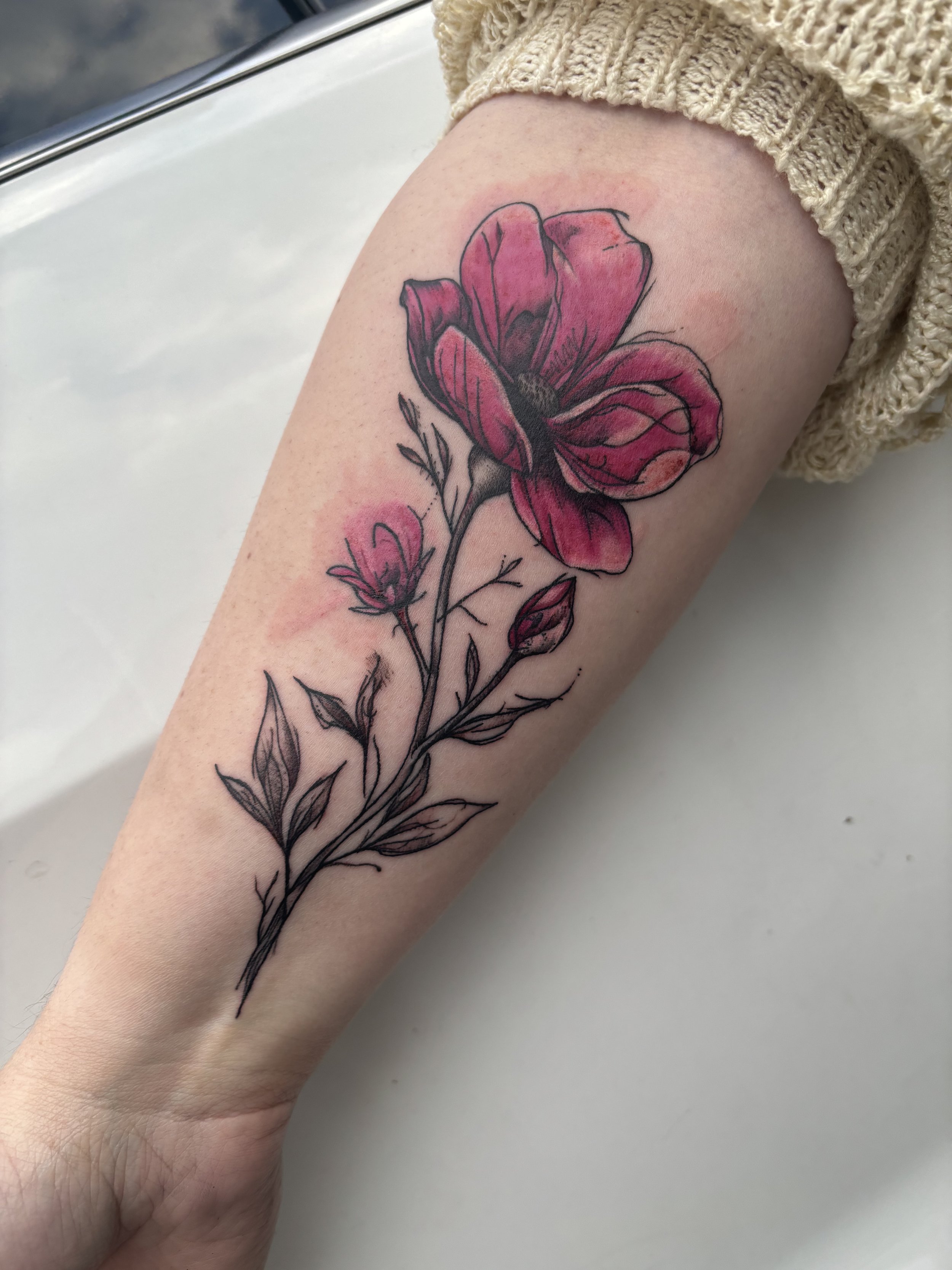 A tattoo of a pink and black flower with leaves on a person's forearm.