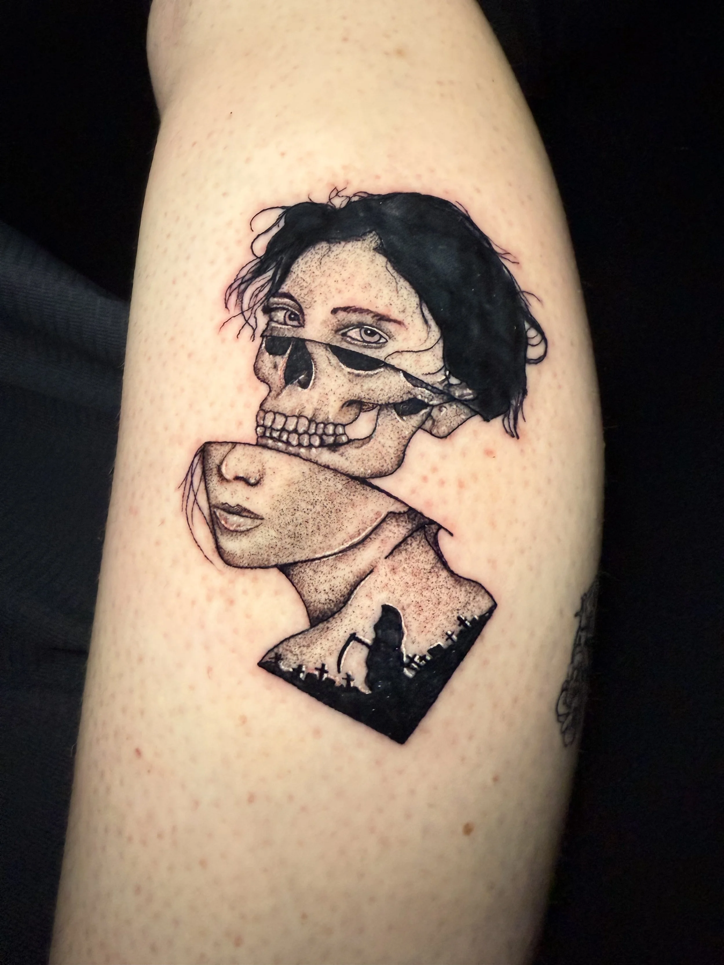 Tattoo of a woman's face partially covered by a skull, with a silhouette of a person and a graveyard scene at the bottom.