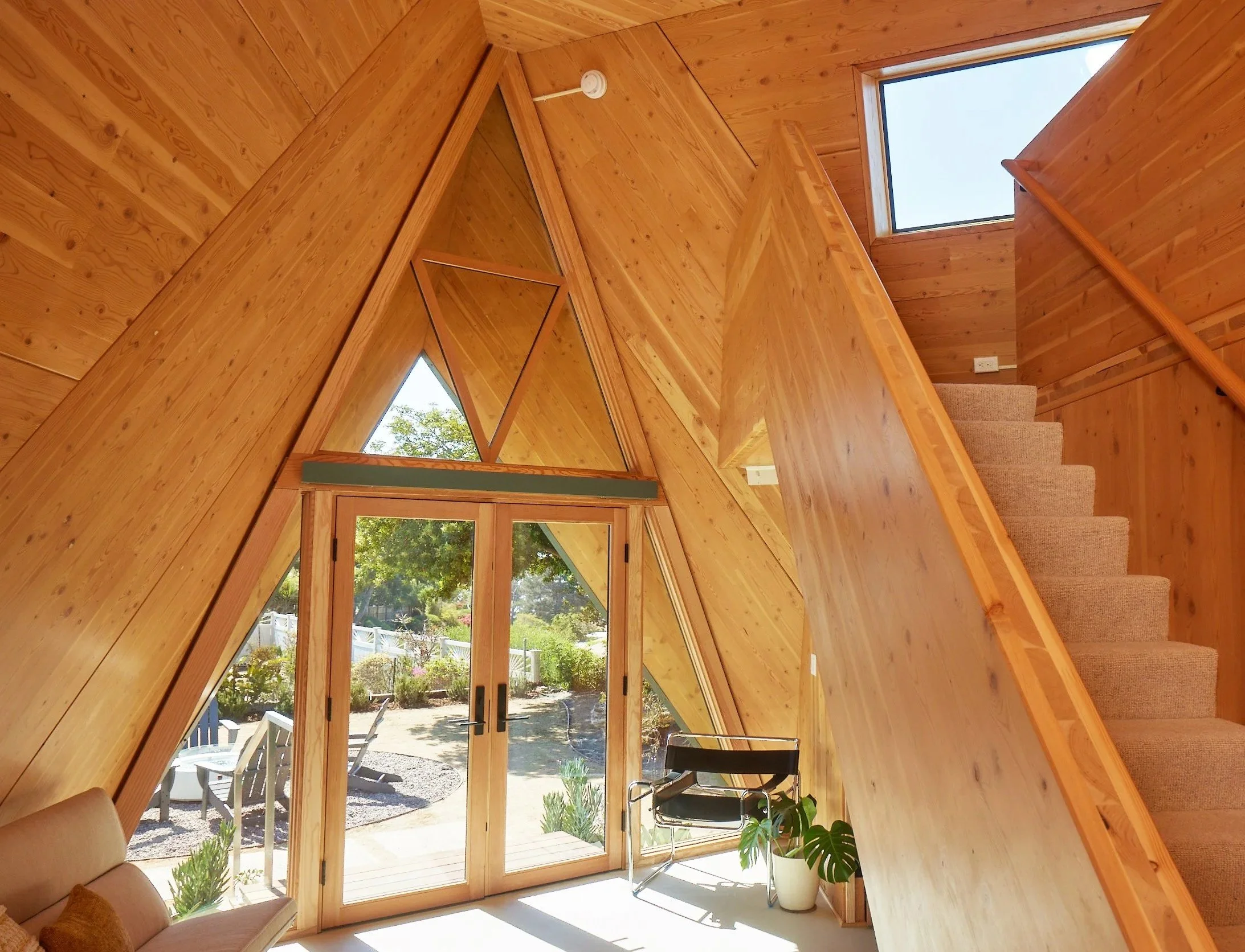 Interior of a wood-paneled house with a staircase leading upstairs, large triangular windows, and a door leading to a garden area outside.