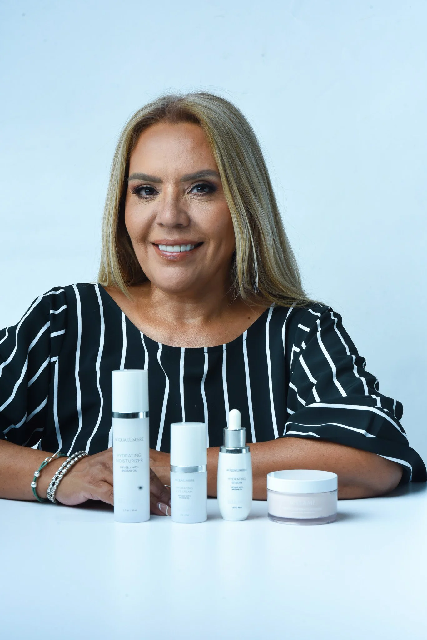A woman with blonde hair and a black and white striped top smiling, sitting at a white table with skincare products including moisturizer, serum, and cream in front of her.