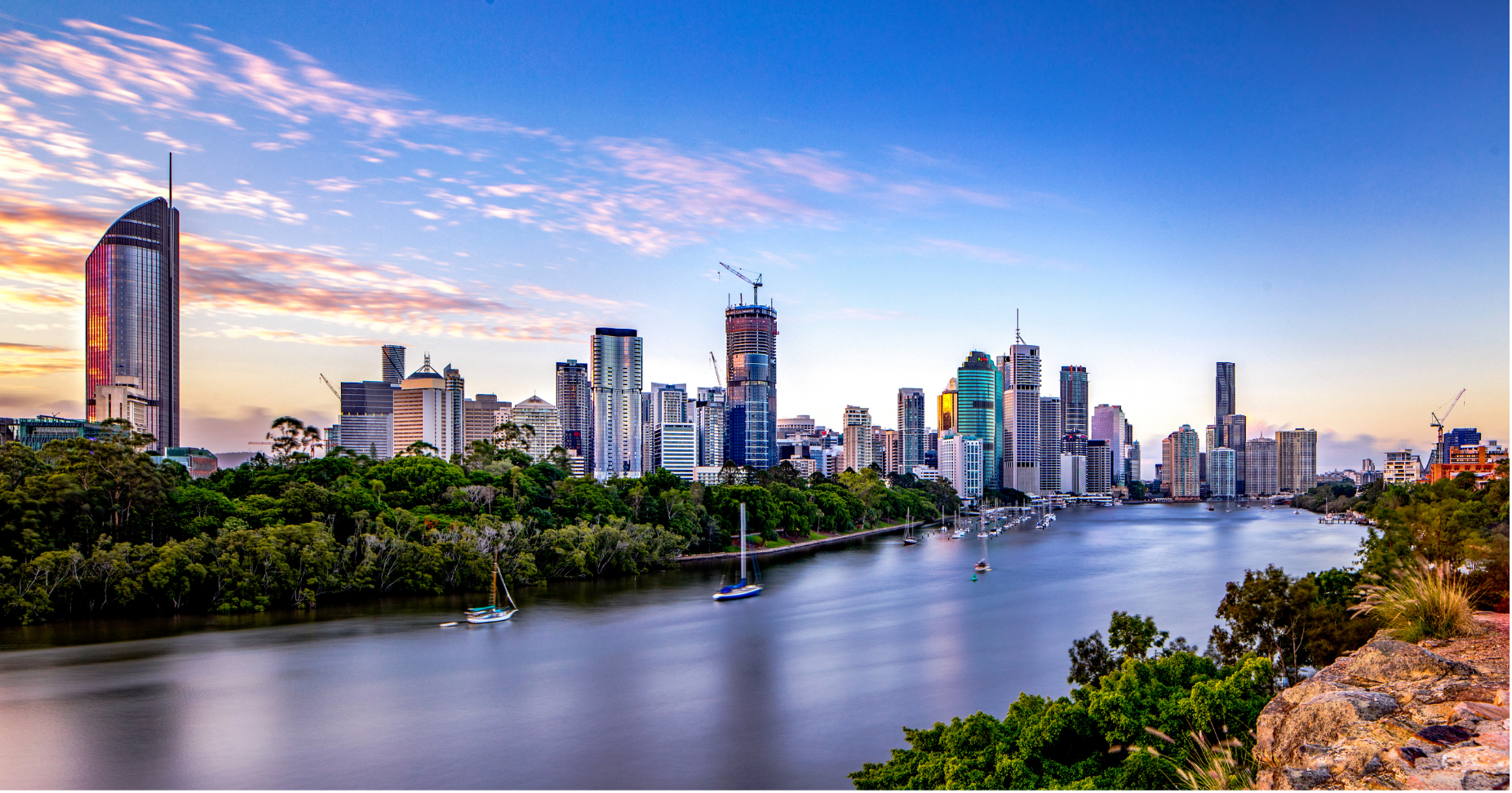 Skyline of Brisbane, Australia, with tall buildings, skyscrapers, a river with sailboats, and greenery in the foreground during sunset.