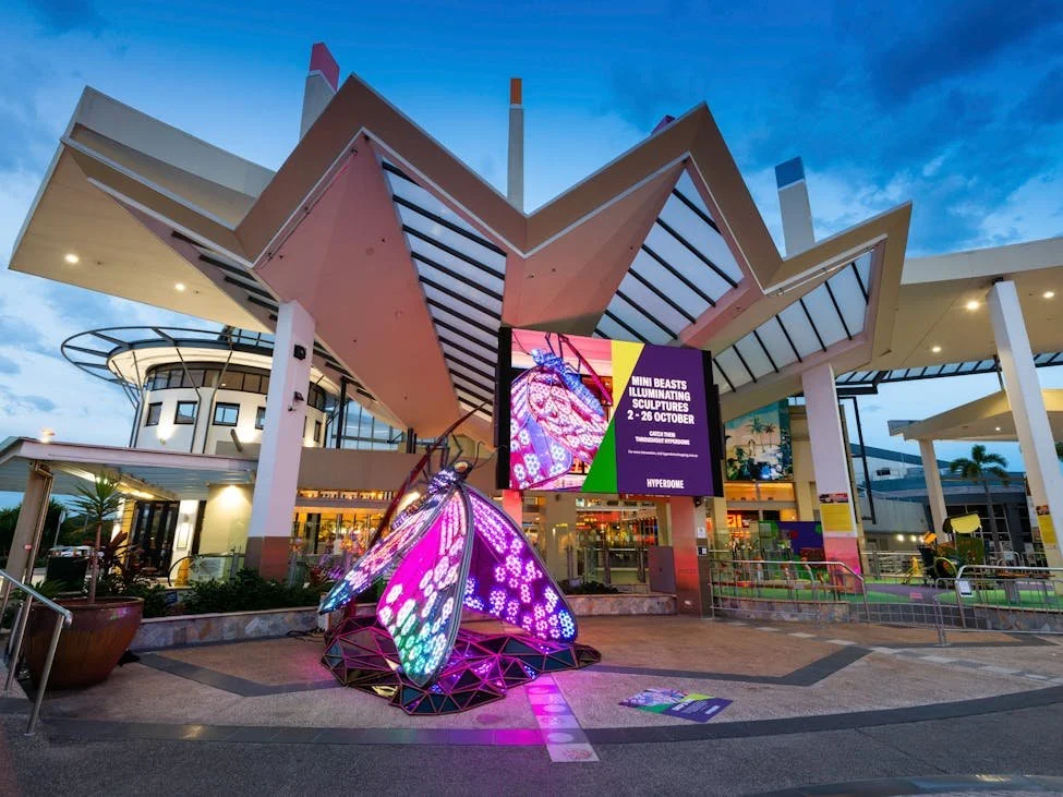 The exterior of a modern building with a geometric roof and large digital screen displaying event information, in an outdoor shopping area or entertainment complex, illuminated by colorful lights with a decorative glowing butterfly sculpture in the foreground.