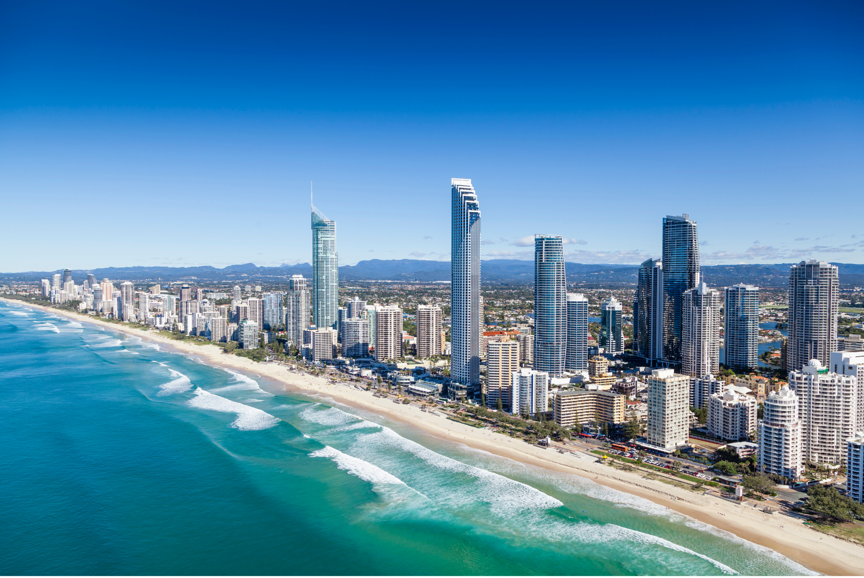 Aerial view of a coastal city with high-rise buildings along a sandy beach, turquoise ocean waves, and mountains in the background under a clear blue sky.