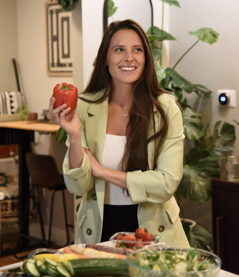 A woman with long brown hair, wearing a light yellow blazer over a white top, smiling and holding a red bell pepper in a kitchen with green plants and a wooden table with vegetables.