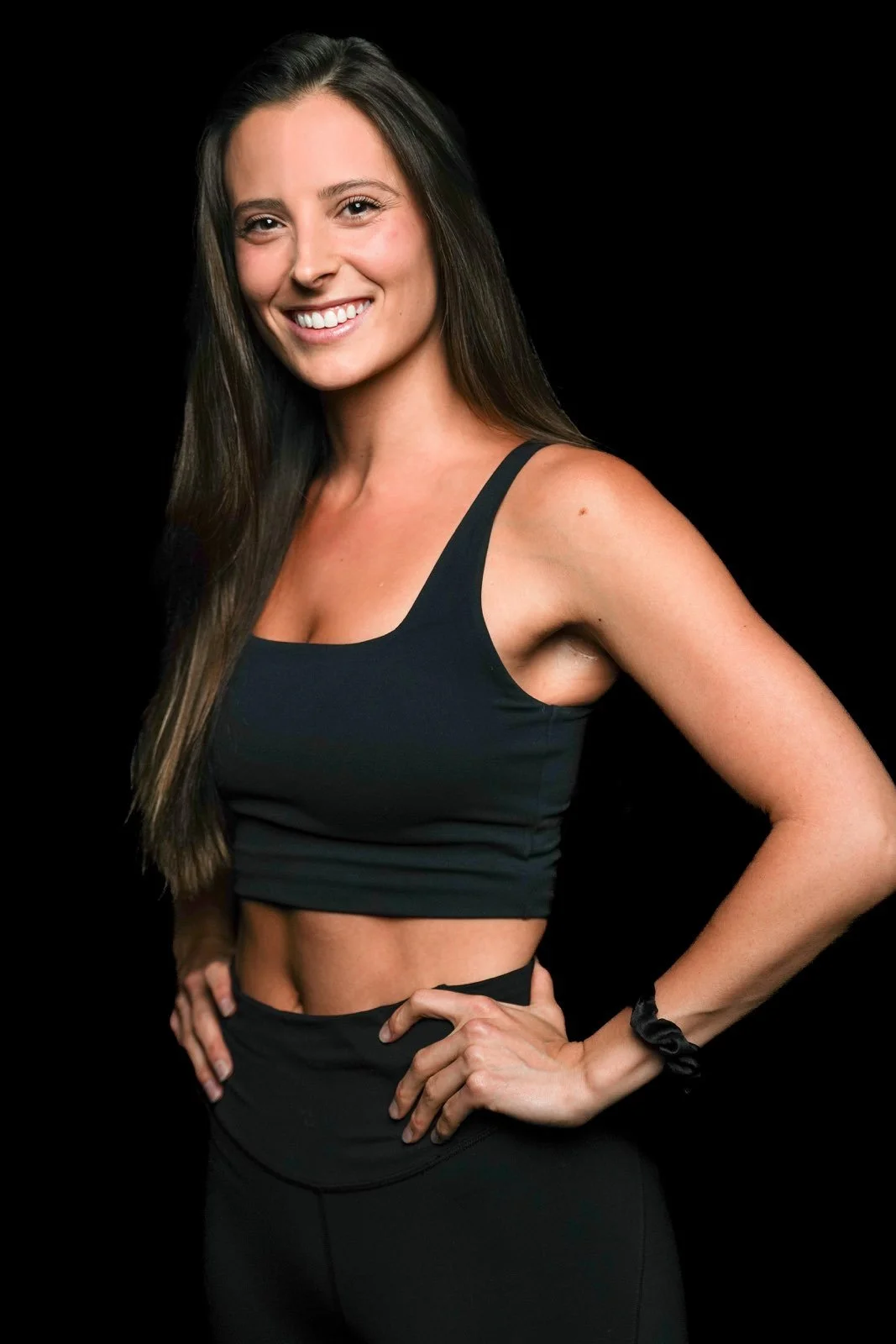 A woman smiling, wearing a black sports bra and leggings, posed with one hand on her hip against a black background.