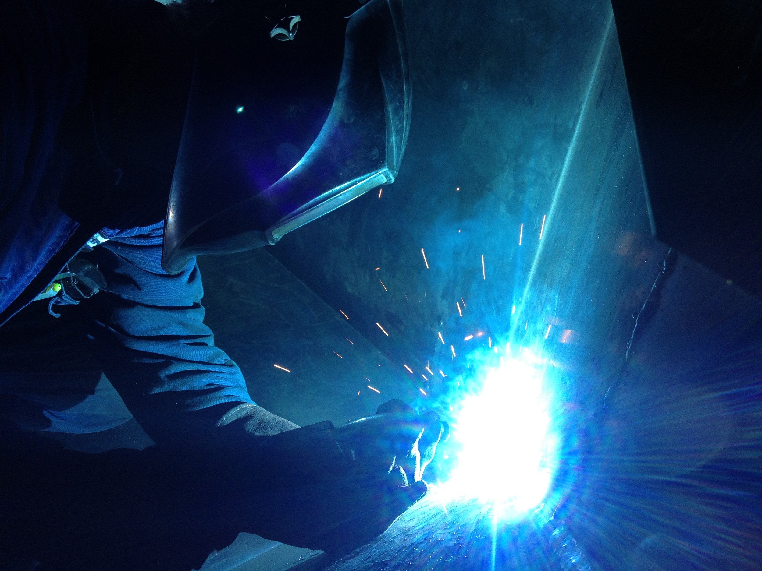 Welder wearing protective gear welding metal, sparks and bright blue light in dark environment.