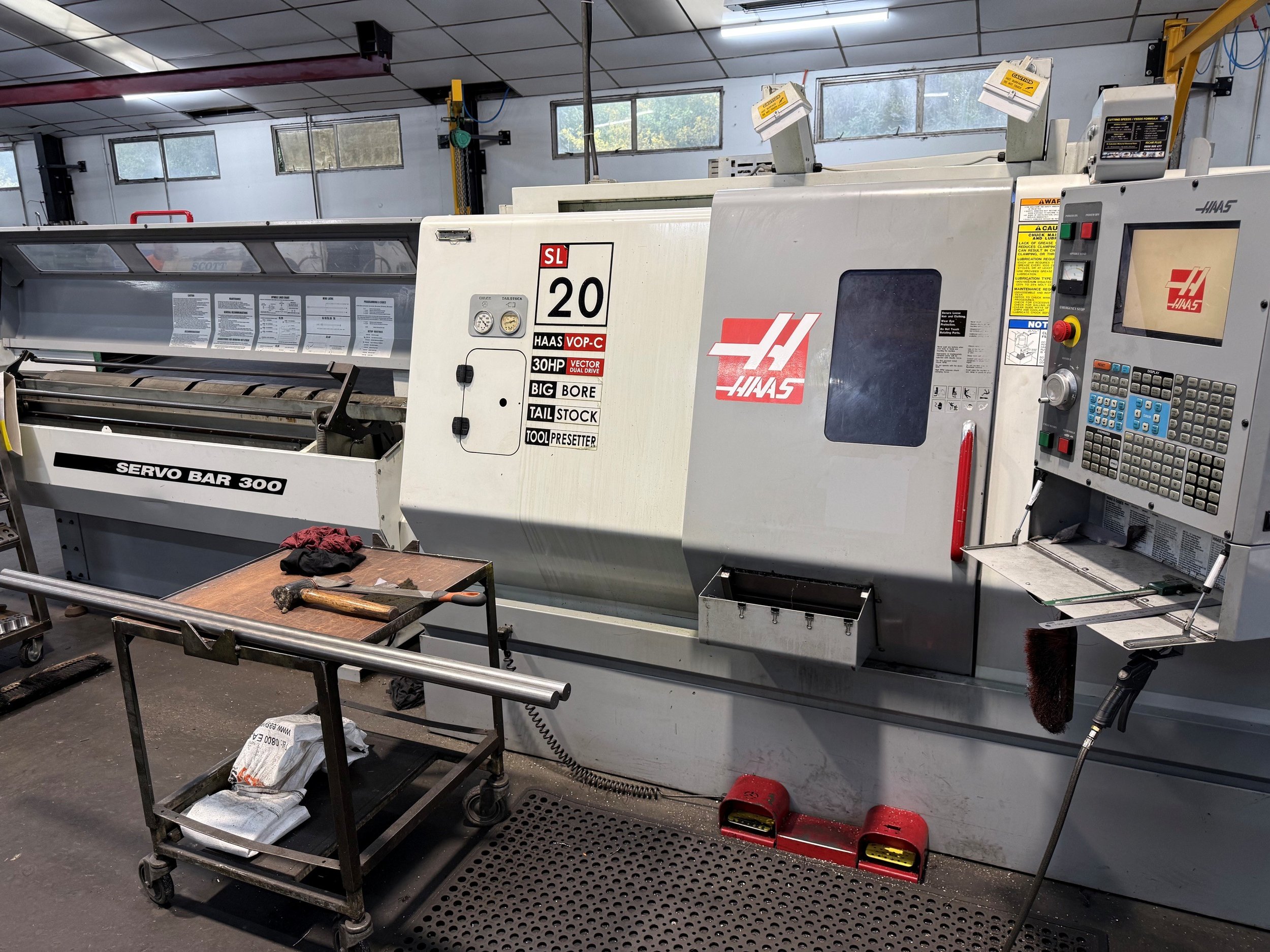 An industrial workshop with large machinery, overhead lighting, and a concrete floor. Multiple pieces of equipment are visible, including a white machine in the foreground labeled 'Servo Bar 300,' with various other machines and tools in the background.