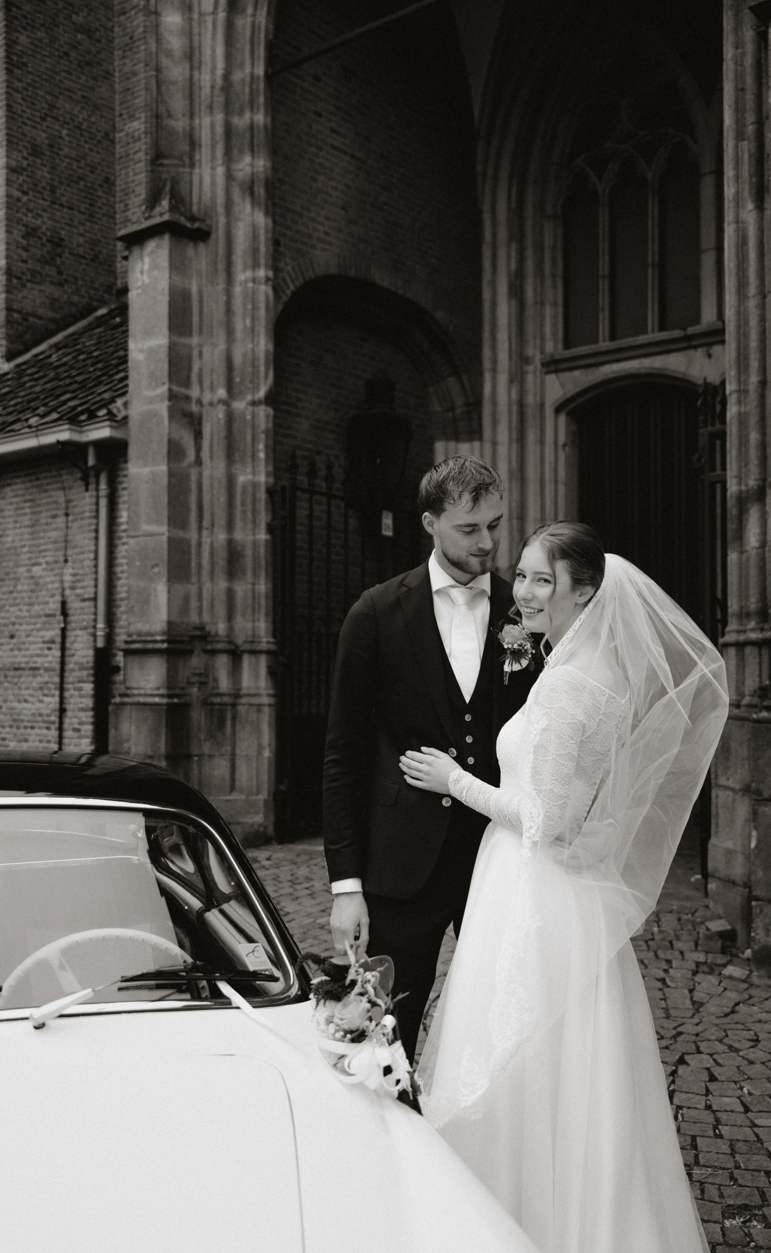 Zwart-wit foto van een bruid en bruidegom in huwelijkskleding voor een oude kerk, lachend en in elkaars gezelschap, met een witte auto als decoratie.