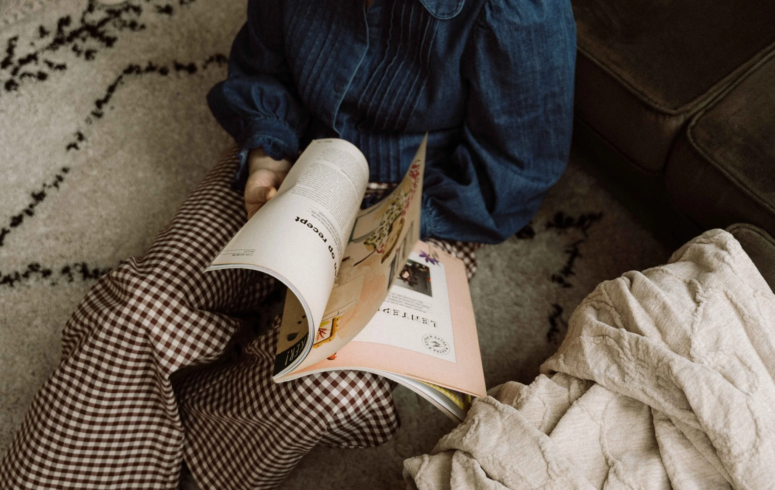 Kind leest een magazine terwijl iemand anders naast hen zit met een gevouwen witte deken, op een tapijt met een zwart-wit patroon, in een woonkamer.