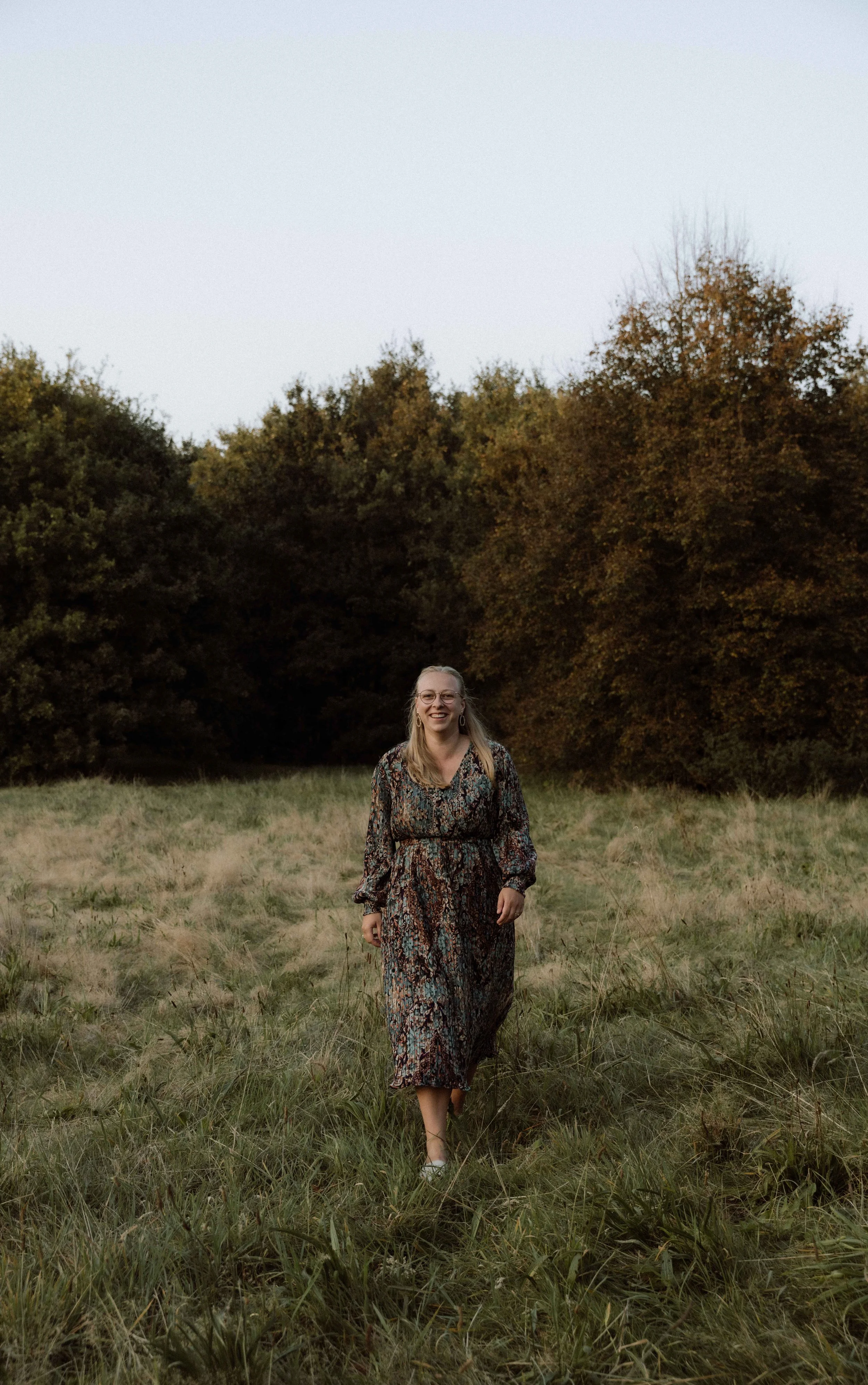 Vrouw in een lange, bloemrijke jurk wandelt door een grasveld met bomen op de achtergrond, bij zonsondergang.