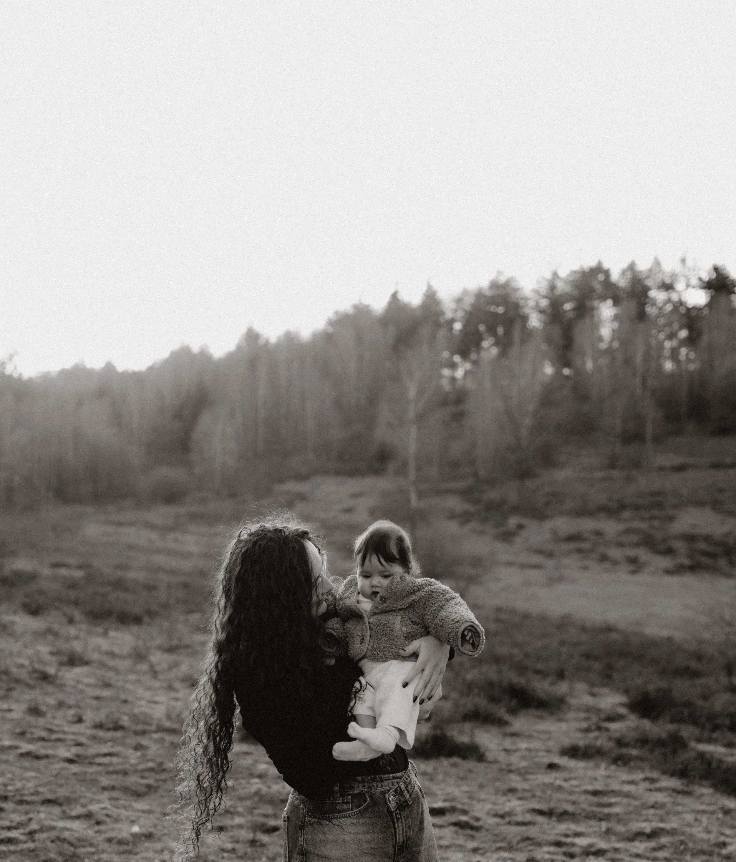 Een vrouw houdt een kind in haar armen in een open veld met een bos op de achtergrond, in zwart-wit.