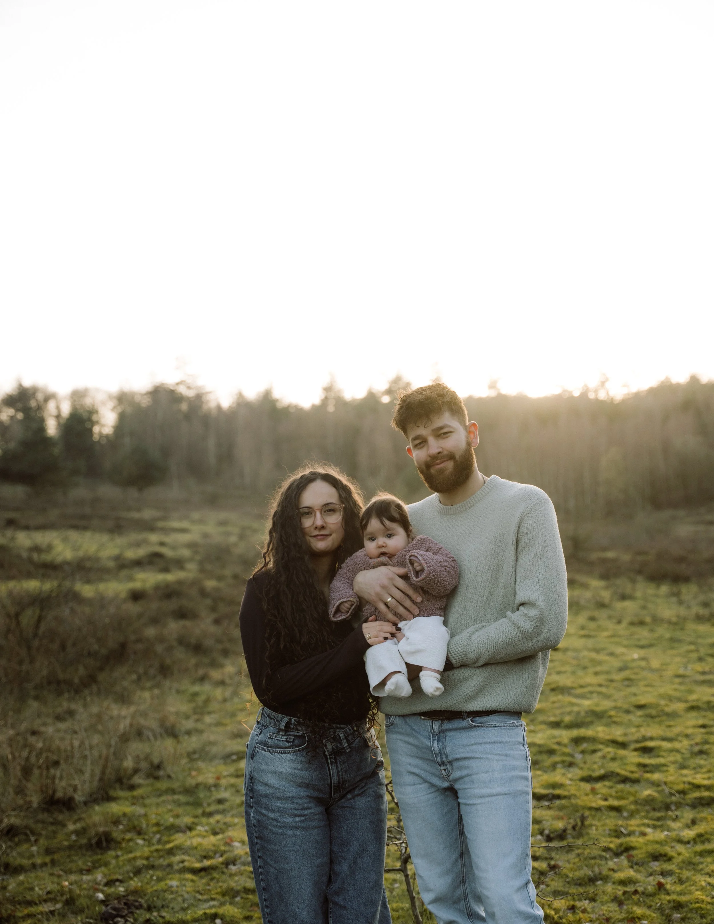 Een gezin bestaande uit een vrouw, man en baby poseert in een groen landschap tijdens zonsondergang.