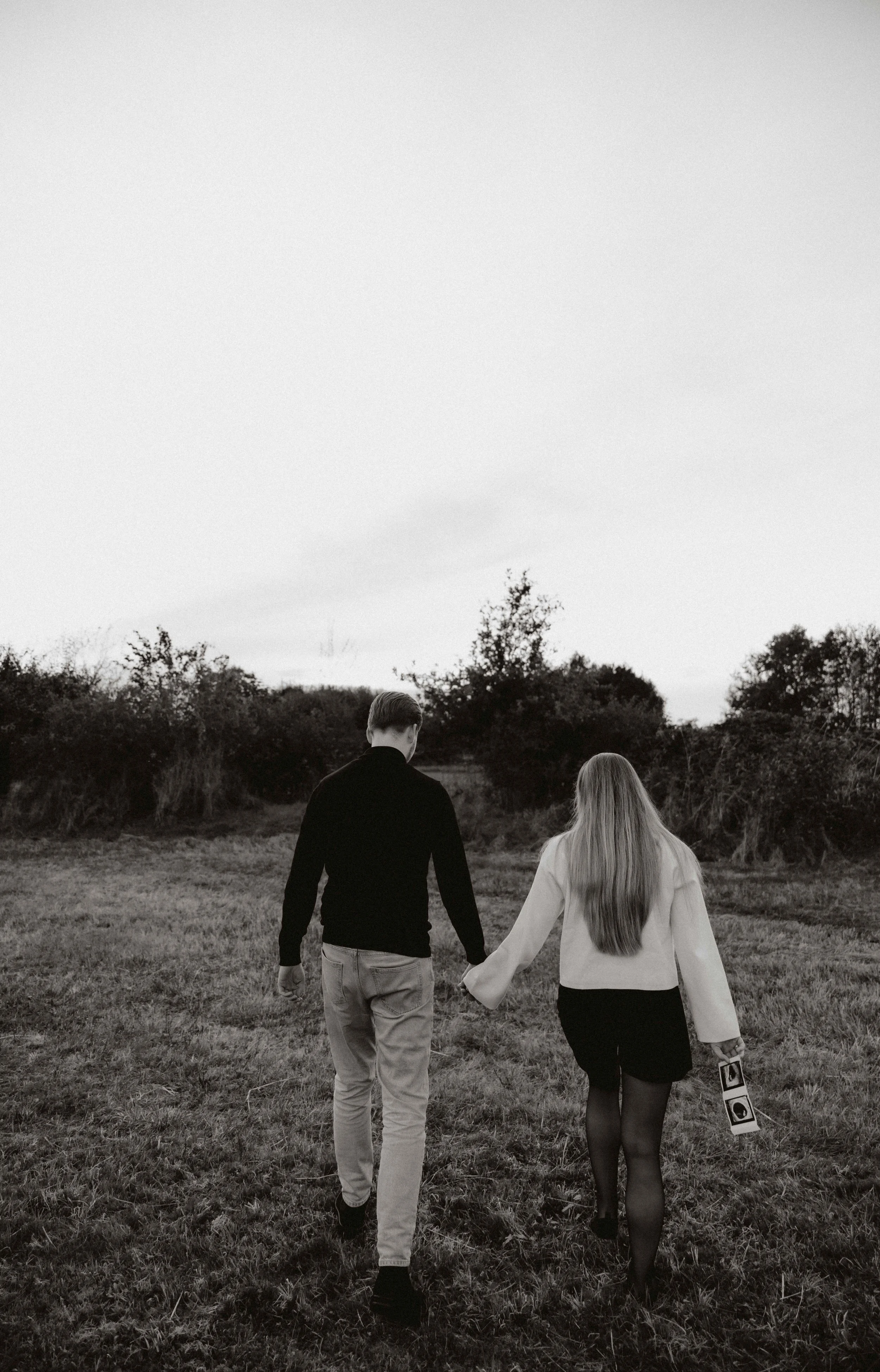 Een zwart-witfoto van een man en een vrouw die samen door een grasveld lopen, hand in hand, met bomen en struiken op de achtergrond.