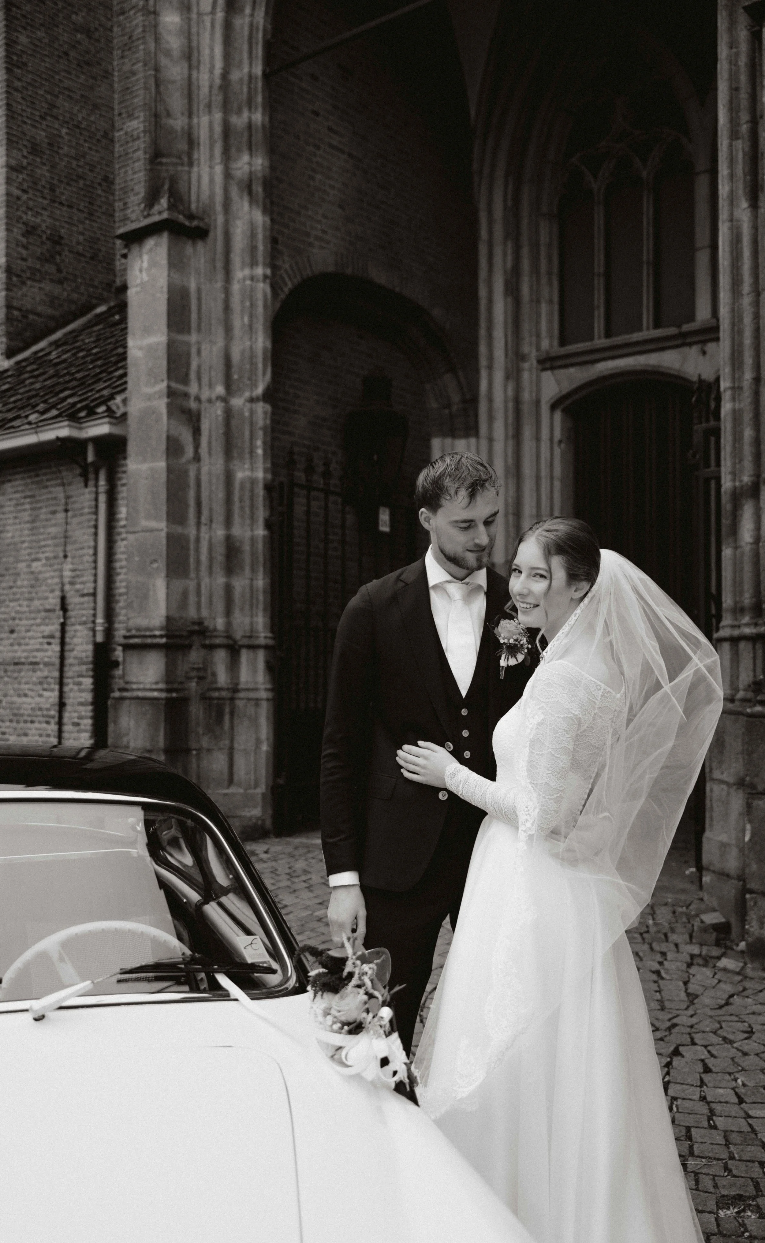 Een zwart-wit foto van een getrouwd stel in formele kleding, met een historische kerk op de achtergrond. Ze staan naast een witte auto versierd met bloemen.