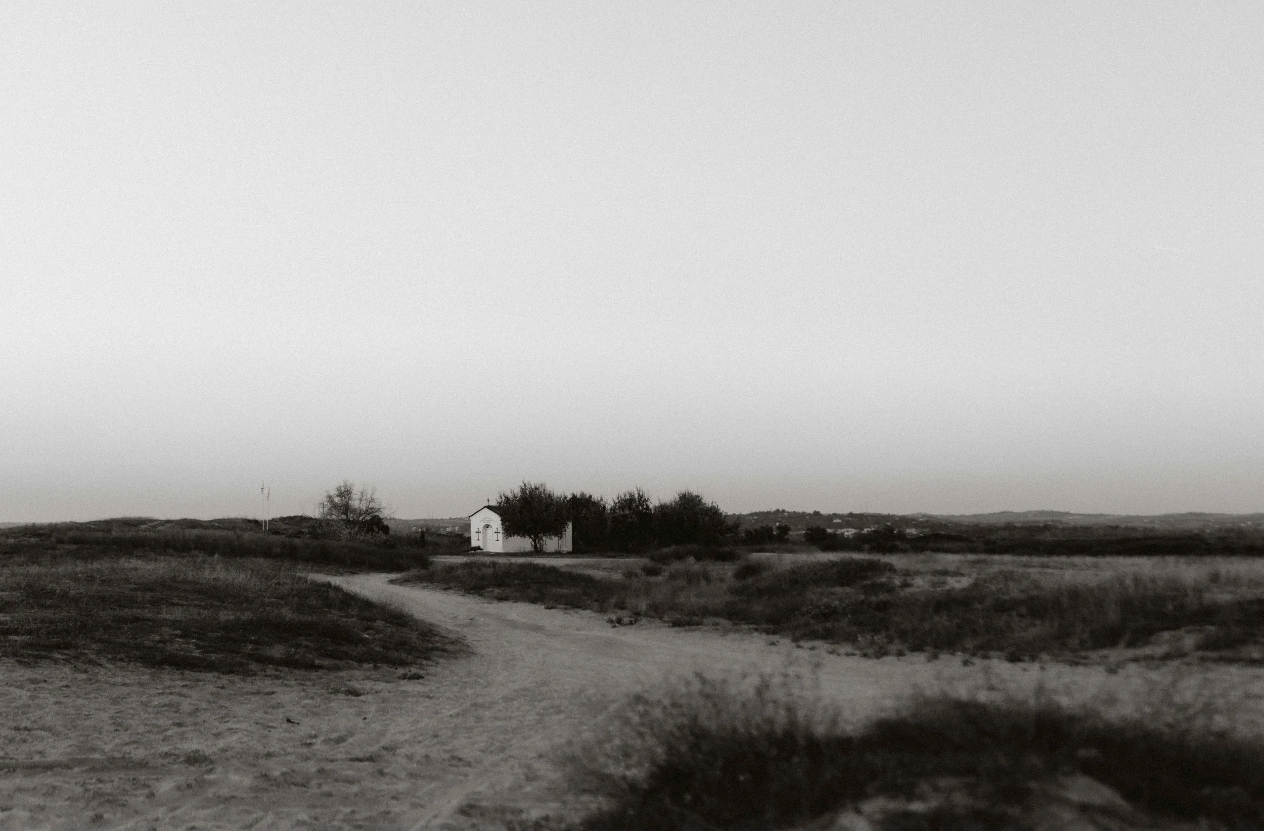 Een zwart-wit landschap met een smalle weg die door een open veld leidt, met een witte kleine kapel en enkele bomen op de achtergrond.