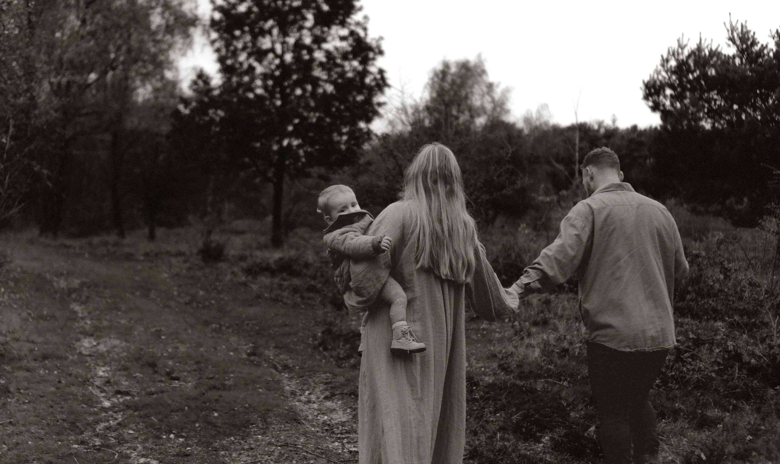 Een familie wandelt op een pad in een bos, de vrouw draagt een kind en de man houdt haar hand vast.
