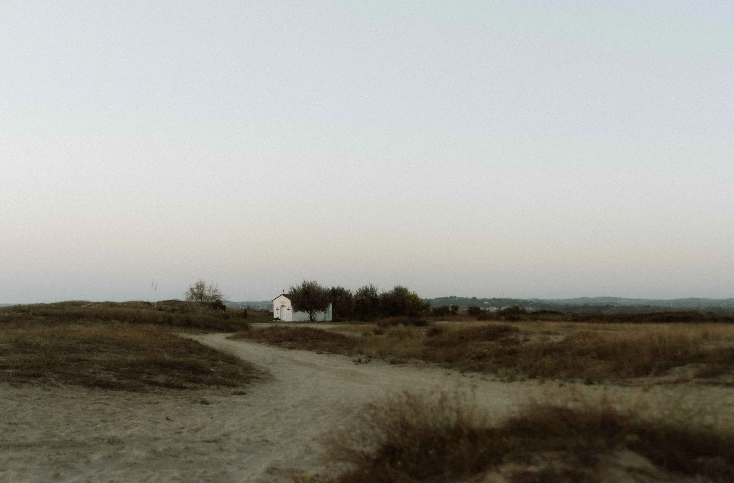Eenzame gekleurde kerk op een zandvlakte met een weg die naar de kerk leidt, omringd door gras en kleine bomen, onder een overwegend heldere hemel.